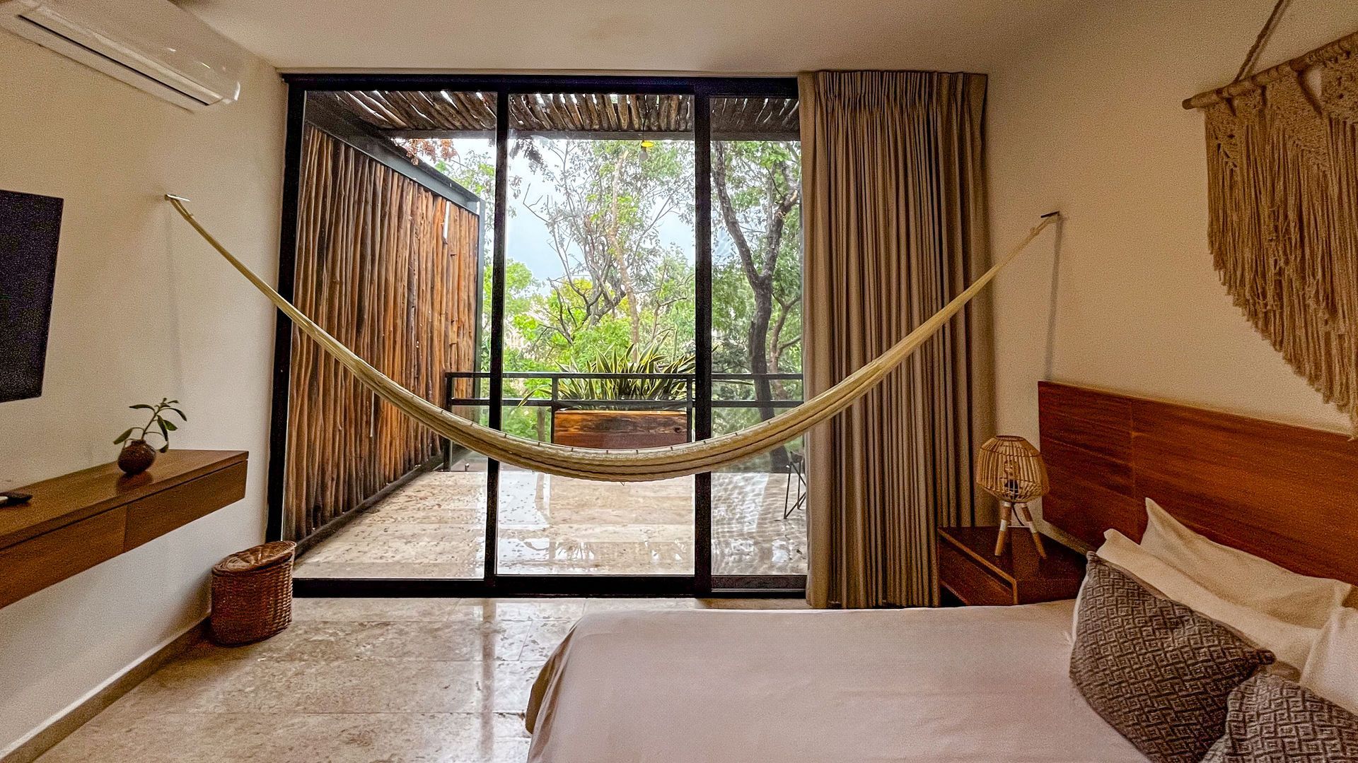 Recámara con hamaca frente a grandes puertas de vidrio que dan a un balcón con detalles en madera, tonos neutros.