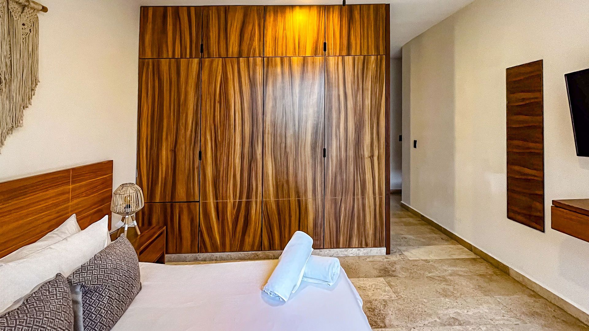 Dormitorio con armario de madera, cama, mesita de noche y televisor. Suelo y paredes de piedra beige.