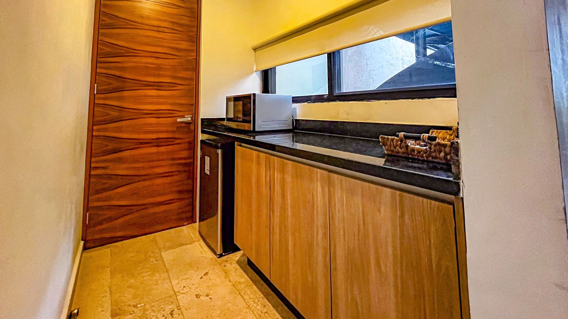 Pequeña cocina con gabinetes de madera, encimera, microondas y mini refrigerador cerca de una ventana y una puerta.