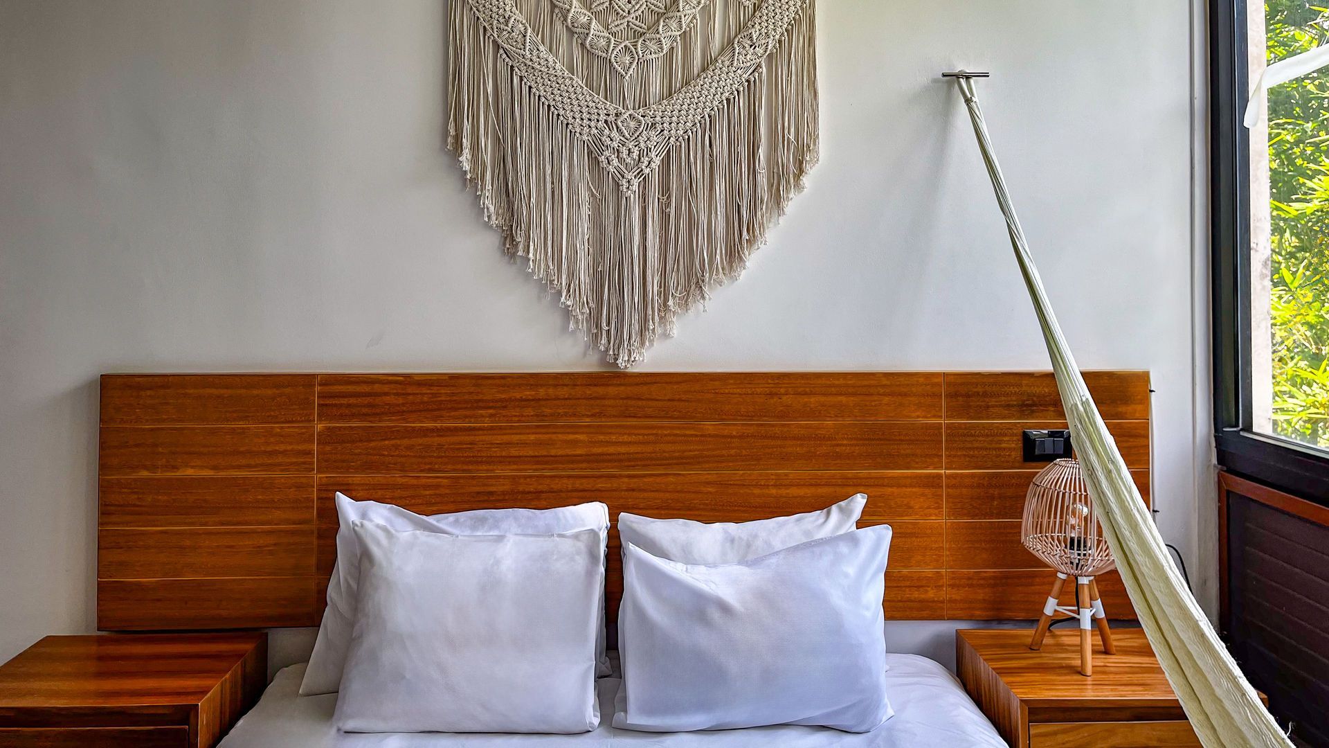 Dormitorio con cabecero de madera, ropa de cama blanca, tapiz de macramé, hamaca y mesitas de noche.