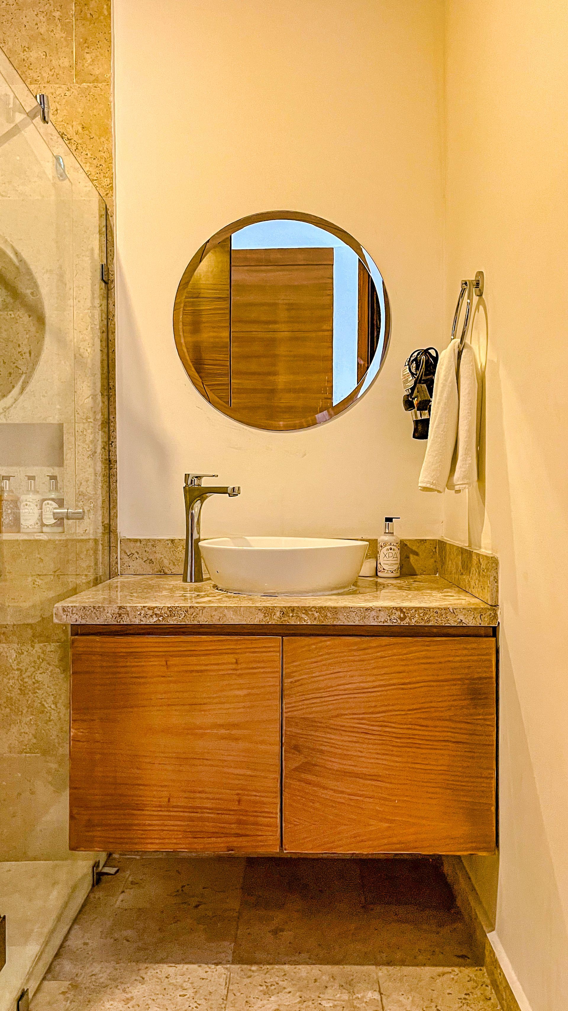 Baño con tocador flotante de madera, espejo redondo y lavabo sobre encimera.