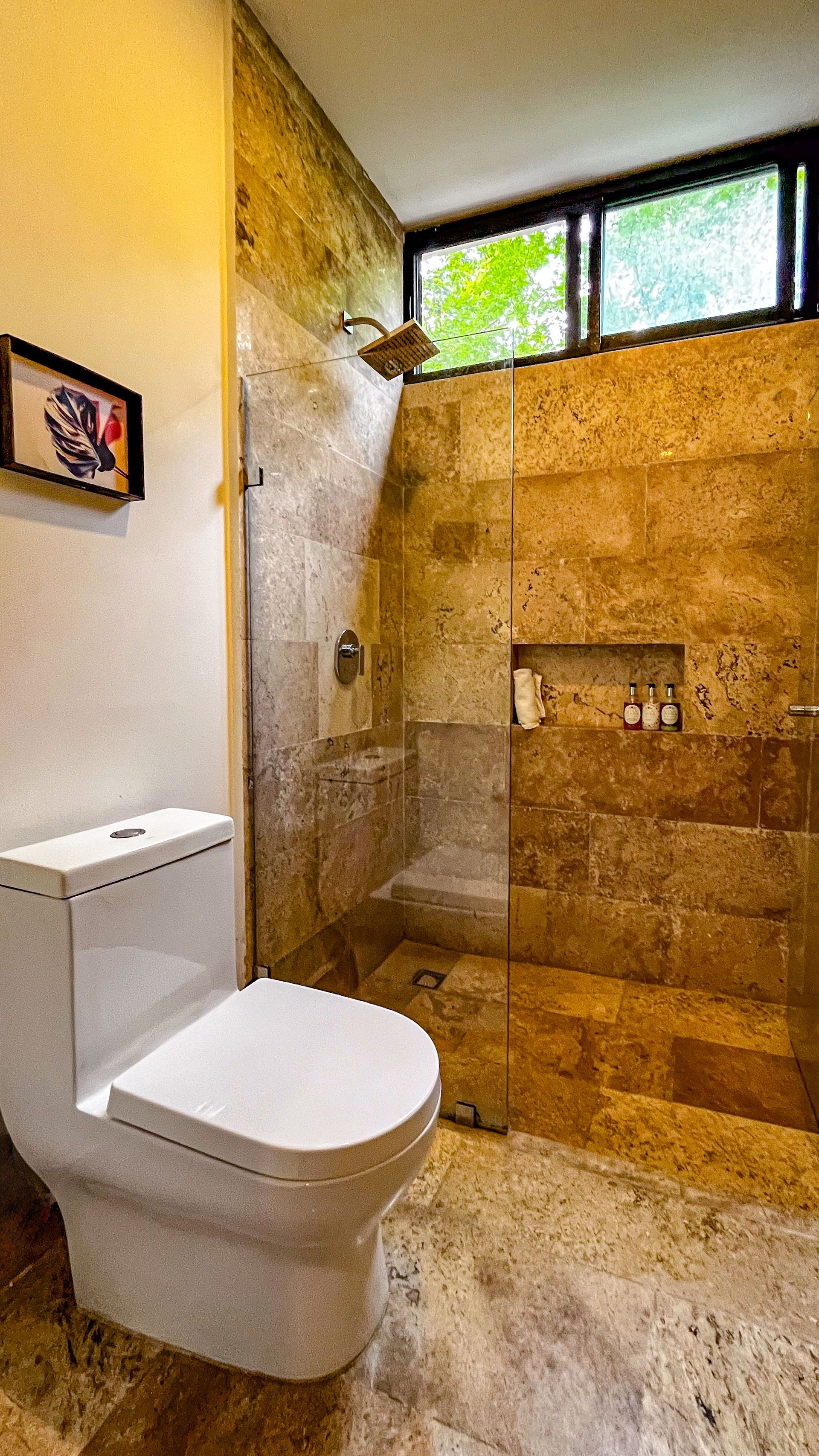 Baño moderno con inodoro blanco, ducha de piedra y una pequeña ventana con vista al follaje verde.