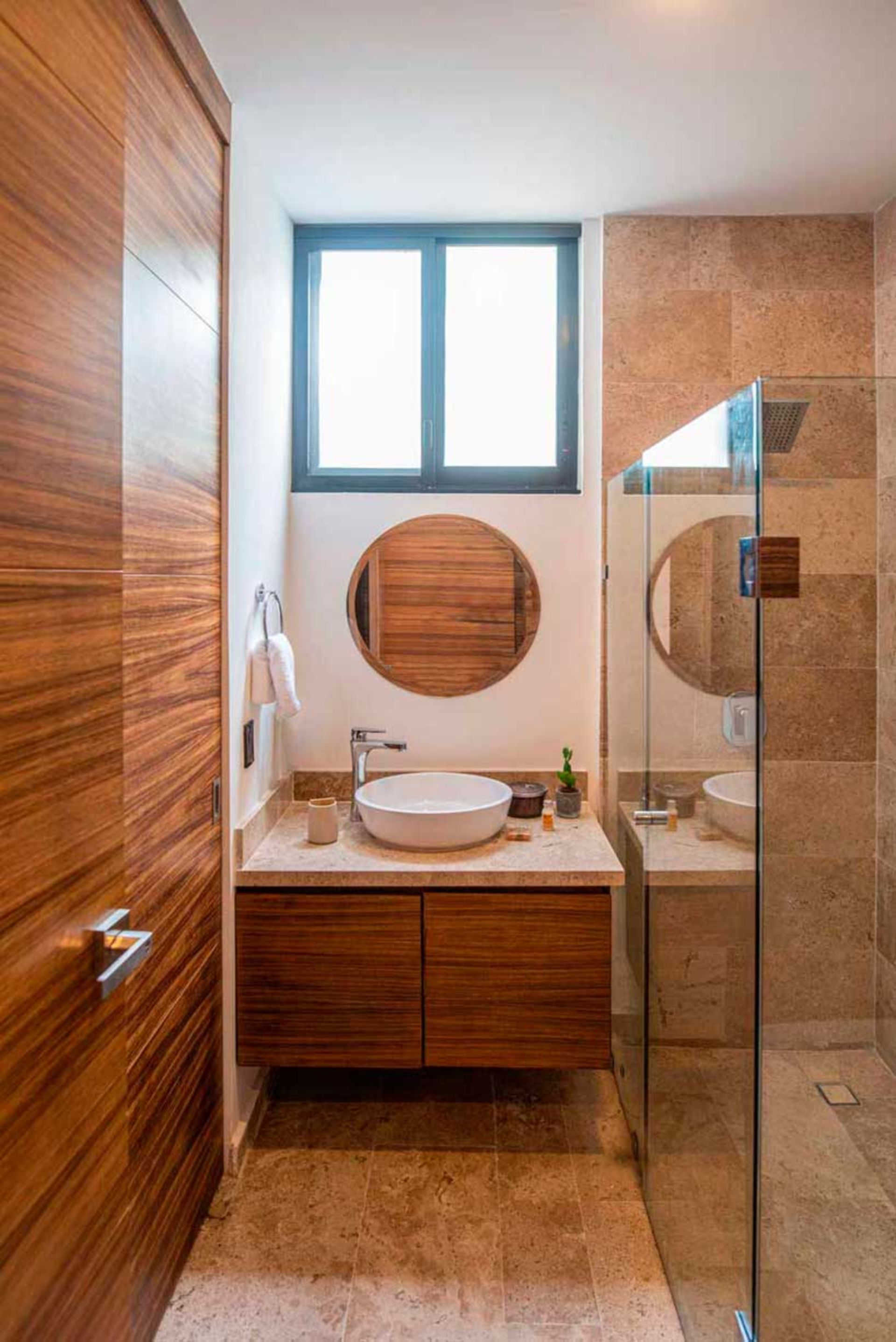 Baño con puerta y tocador de madera, espejo redondo, lavabo blanco y ducha con puertas de cristal.