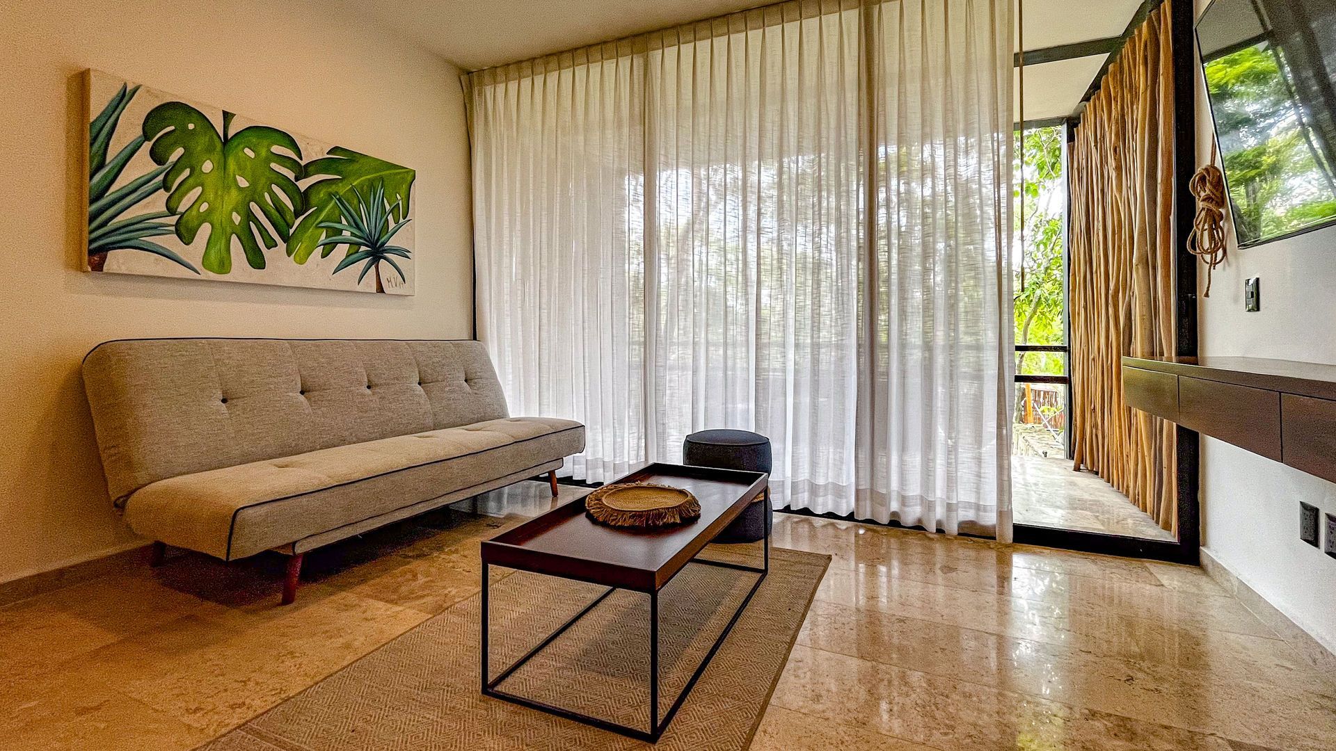 Sala de estar con un sofá beige, arte tropical, cortinas transparentes y una pared con acento de madera.