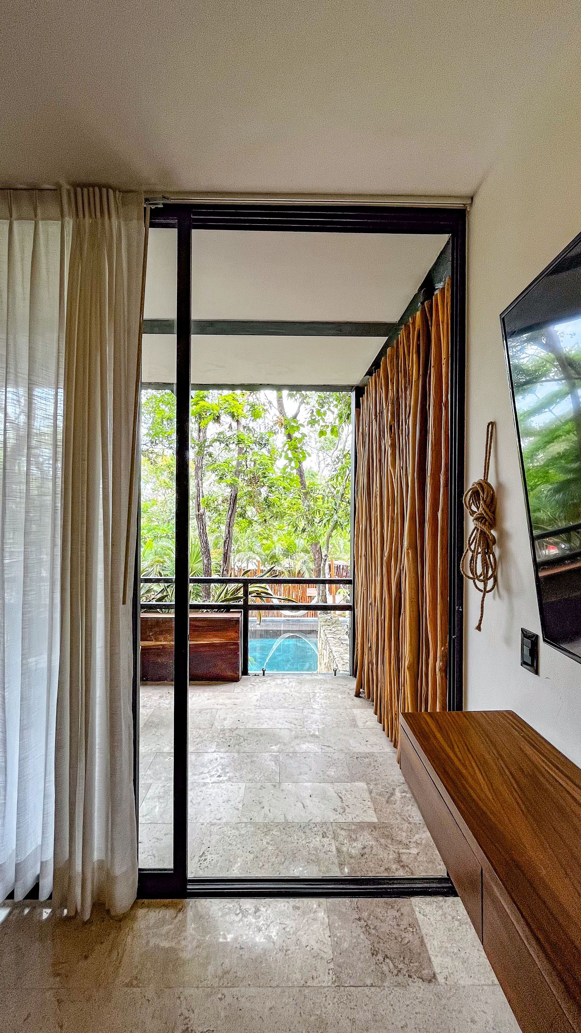 Habitación con balcón con vistas a la piscina. Puerta de cristal, pared con detalles de madera y cortinas beige.
