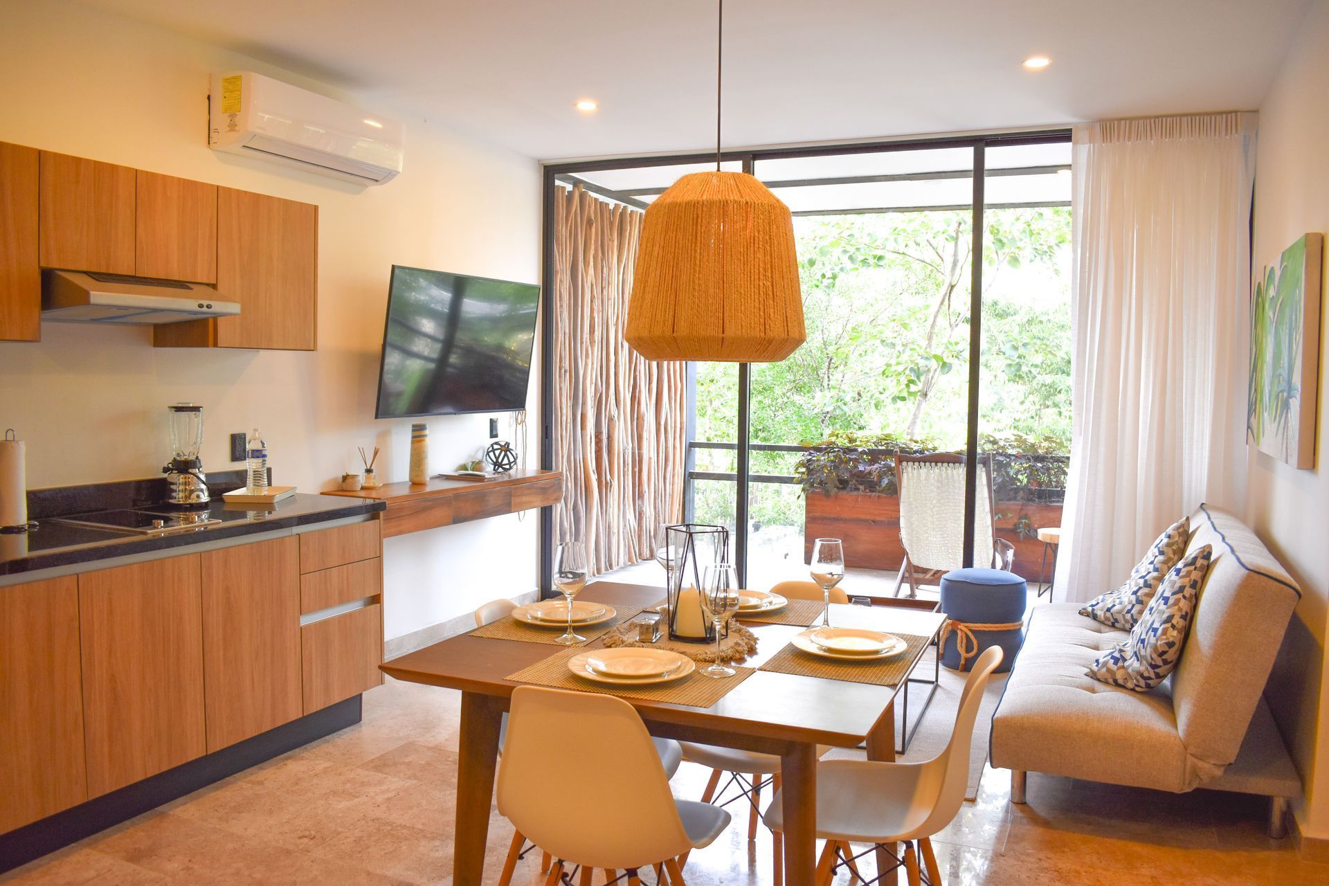 Comedor con cocina: muebles de madera, mesa para cuatro, sofá y balcón con vegetación.