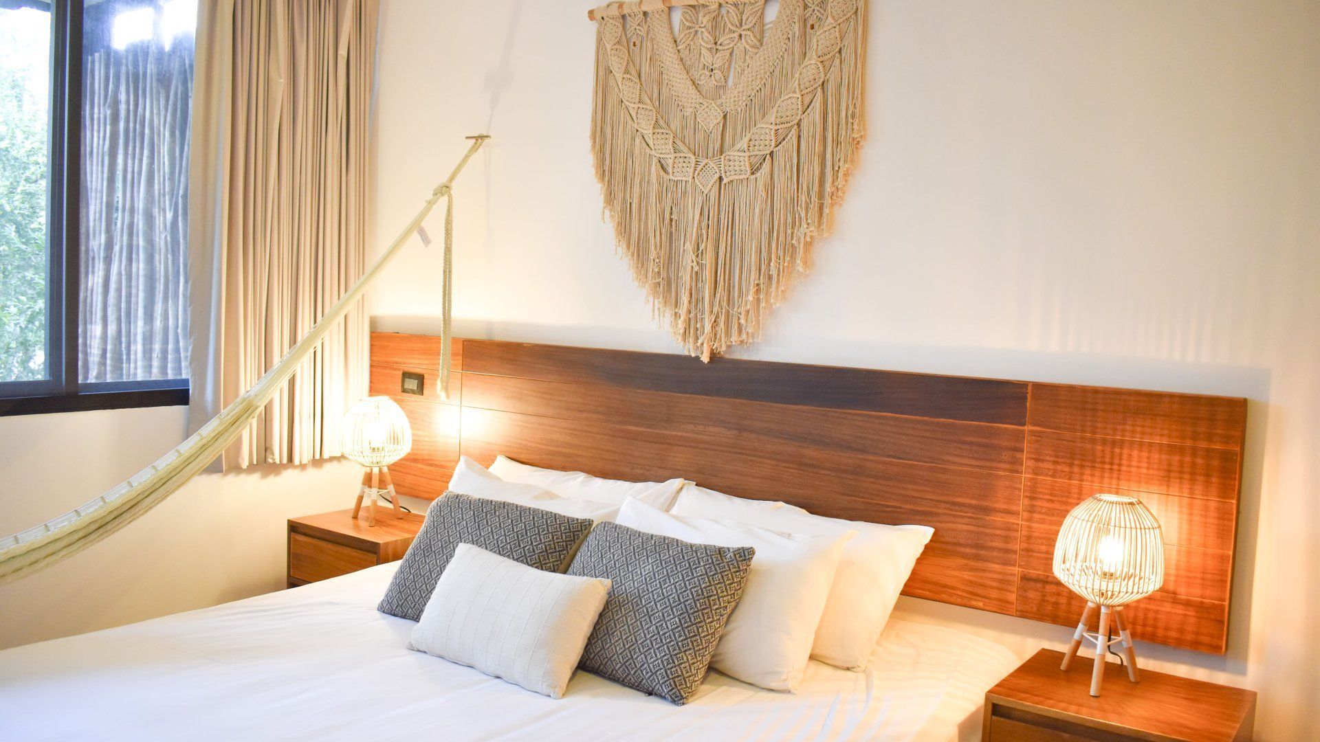 Dormitorio con hamaca, macramé decorativo, cama con almohadas, cabecero de madera y lámparas de noche.