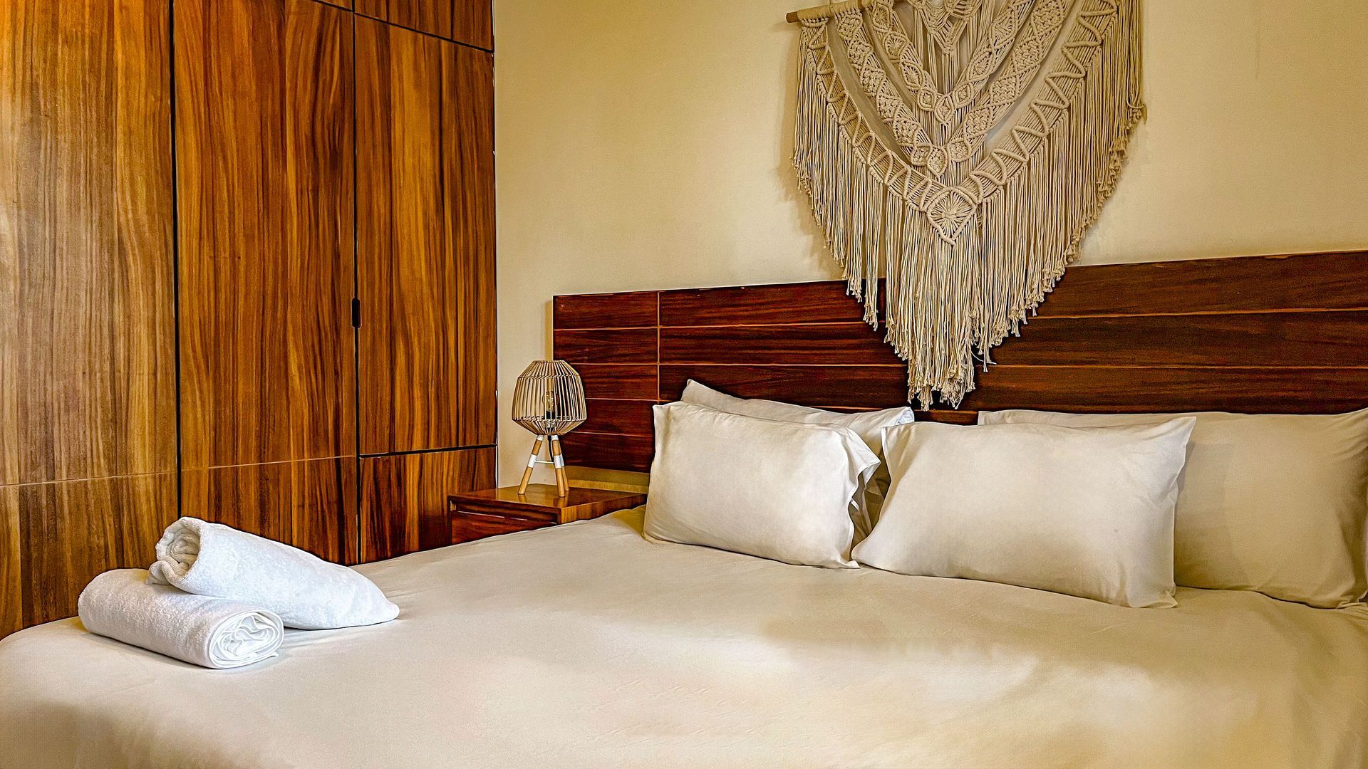 Dormitorio con cabecero de madera y armario, ropa de cama blanca y tapiz de macramé.