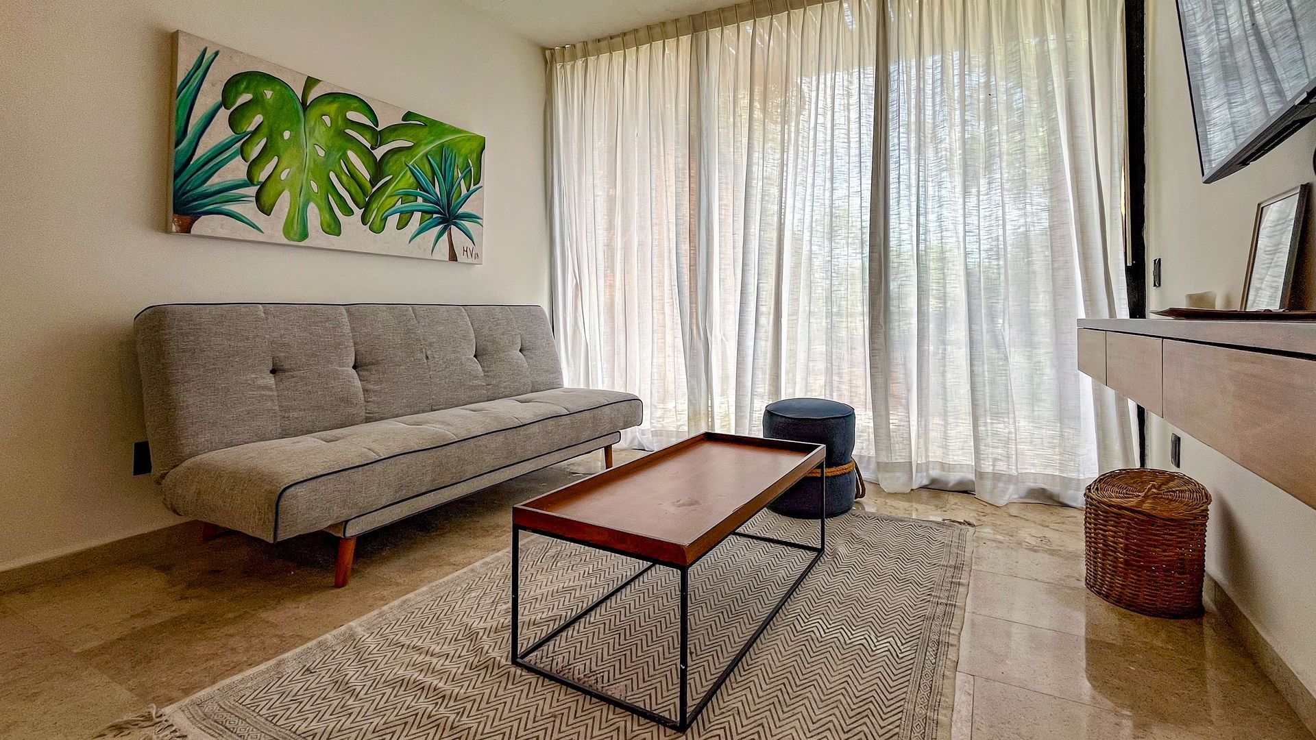 Sala de estar con sofá gris, mesa de café, alfombra, obras de arte y cortinas transparentes.