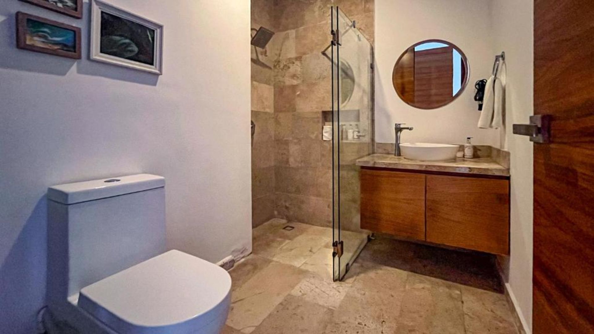 Baño con inodoro, ducha, tocador y obras de arte; tonos neutros y luz natural.