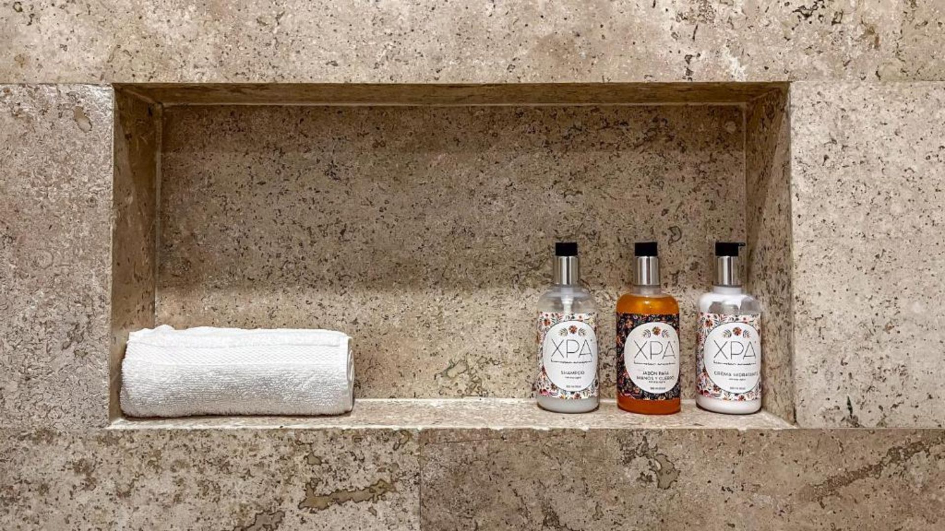 Estante de ducha empotrado con toalla y tres botellas de artículos de tocador. Fondo de piedra marrón claro.