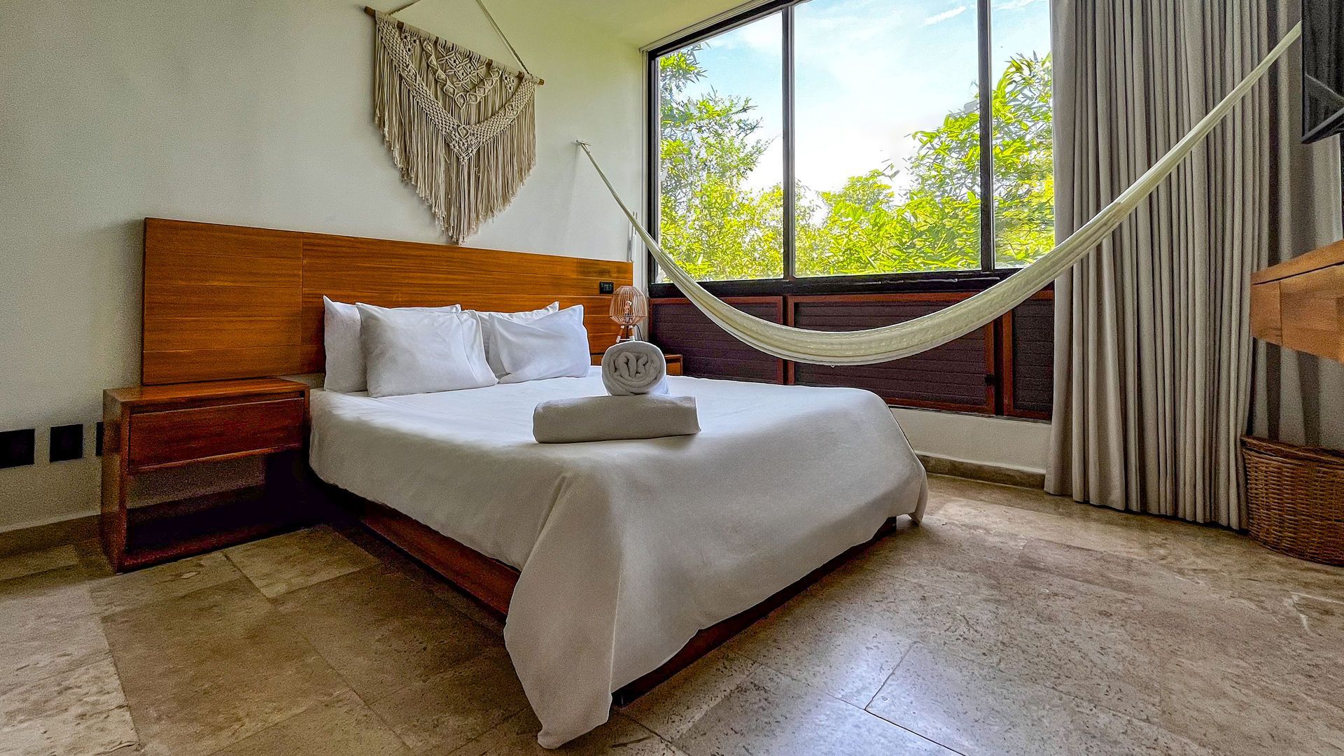 Dormitorio con cama, mesita de noche, ventanal, hamaca y tapiz de macramé.