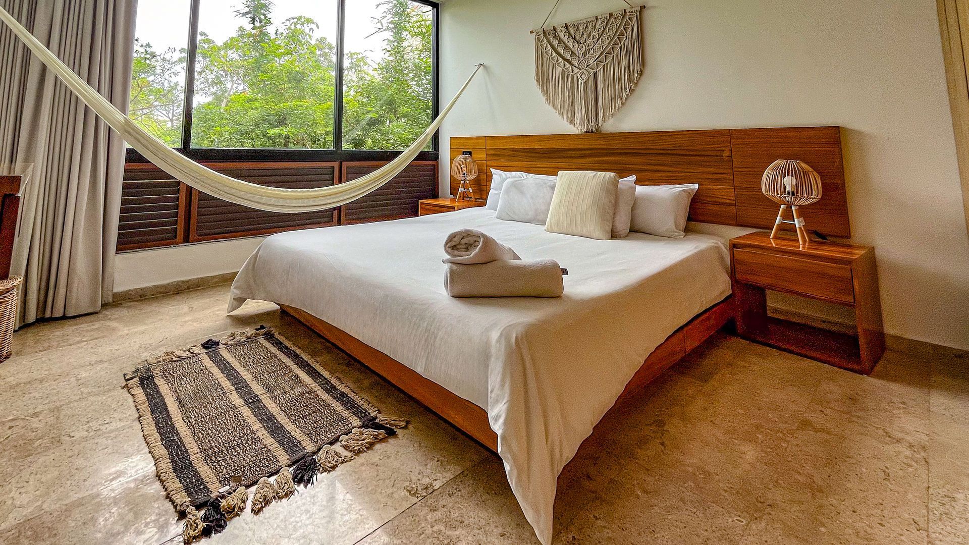 Dormitorio con cama grande, hamaca y ventanal con vista a los árboles. Tonos neutros y luz natural.