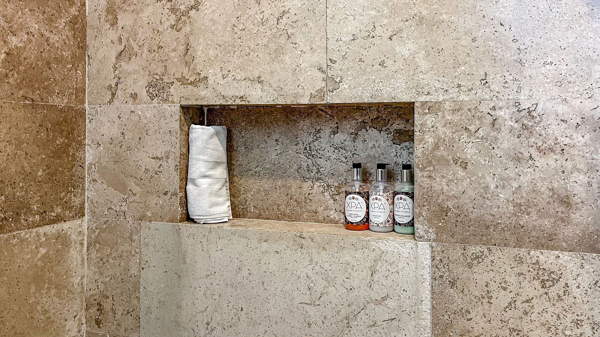 Estante de ducha empotrado con artículos de aseo. Azulejos de piedra beige. Toalla y botellas dentro del hueco.