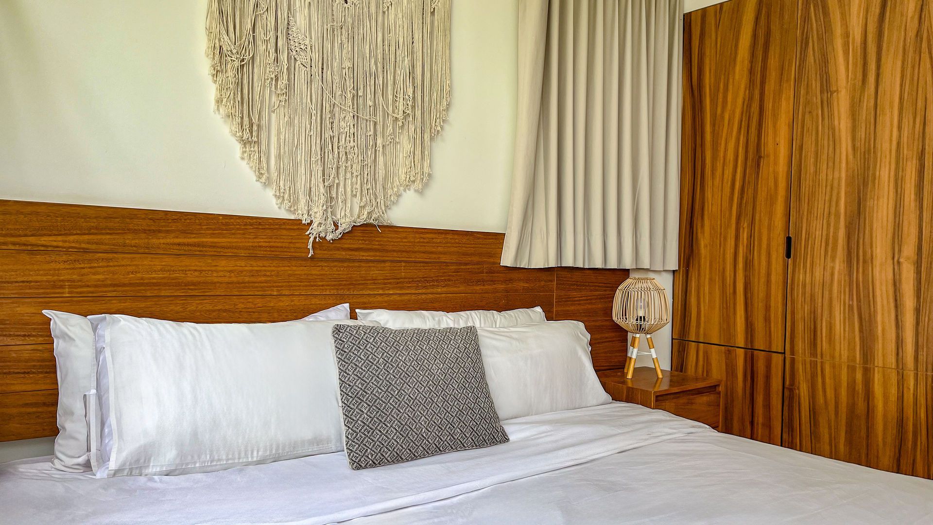 Dormitorio con cabecero de madera, ropa de cama blanca, tapiz de macramé y armario de madera.