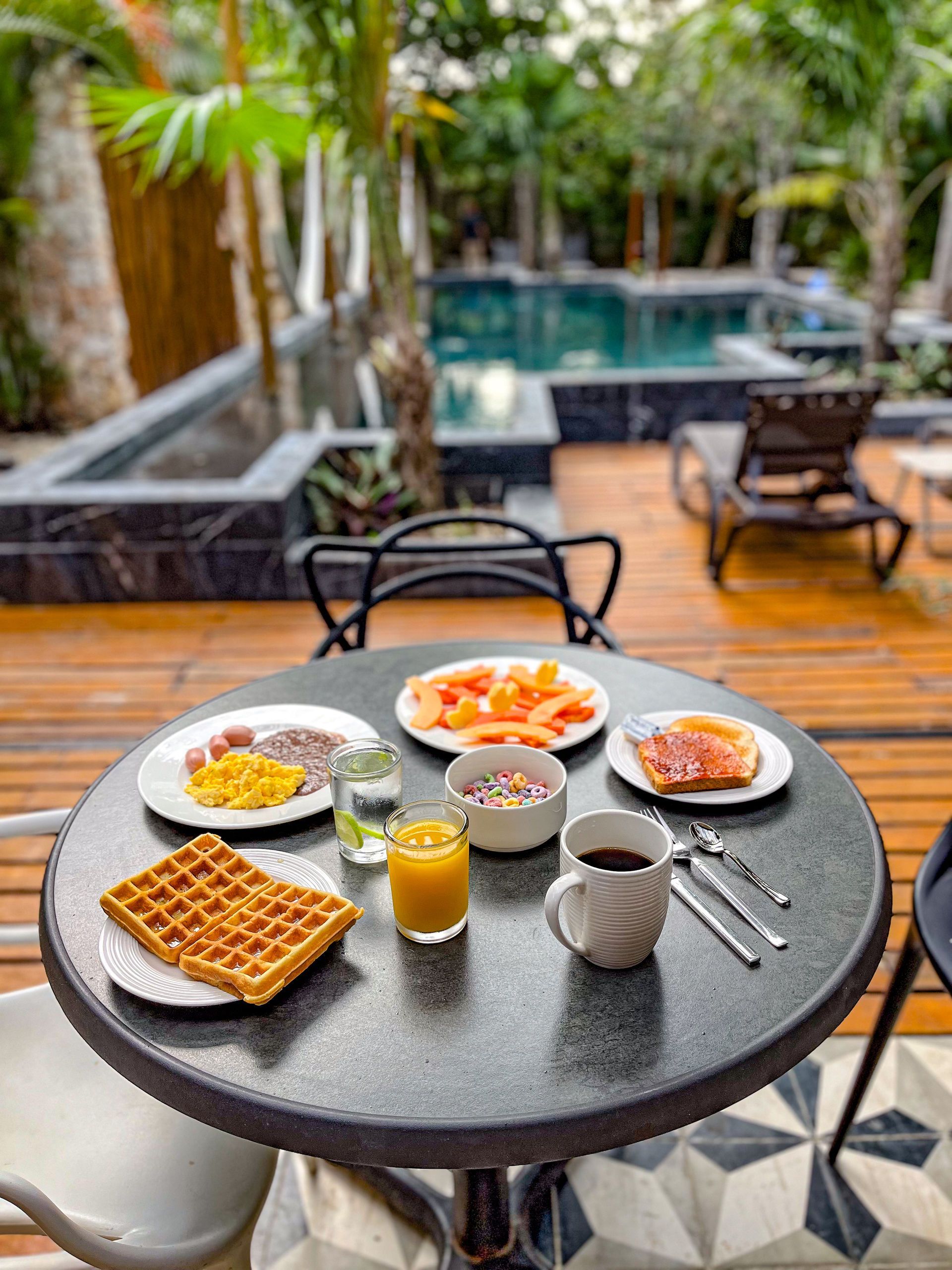 Mesa de desayuno con waffles, fruta y café con vista a una piscina.