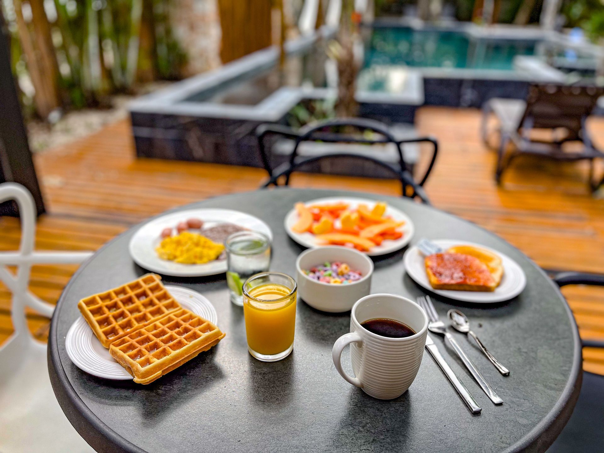Desayuno servido en una mesa redonda al aire libre, waffles, fruta y café junto a una piscina.