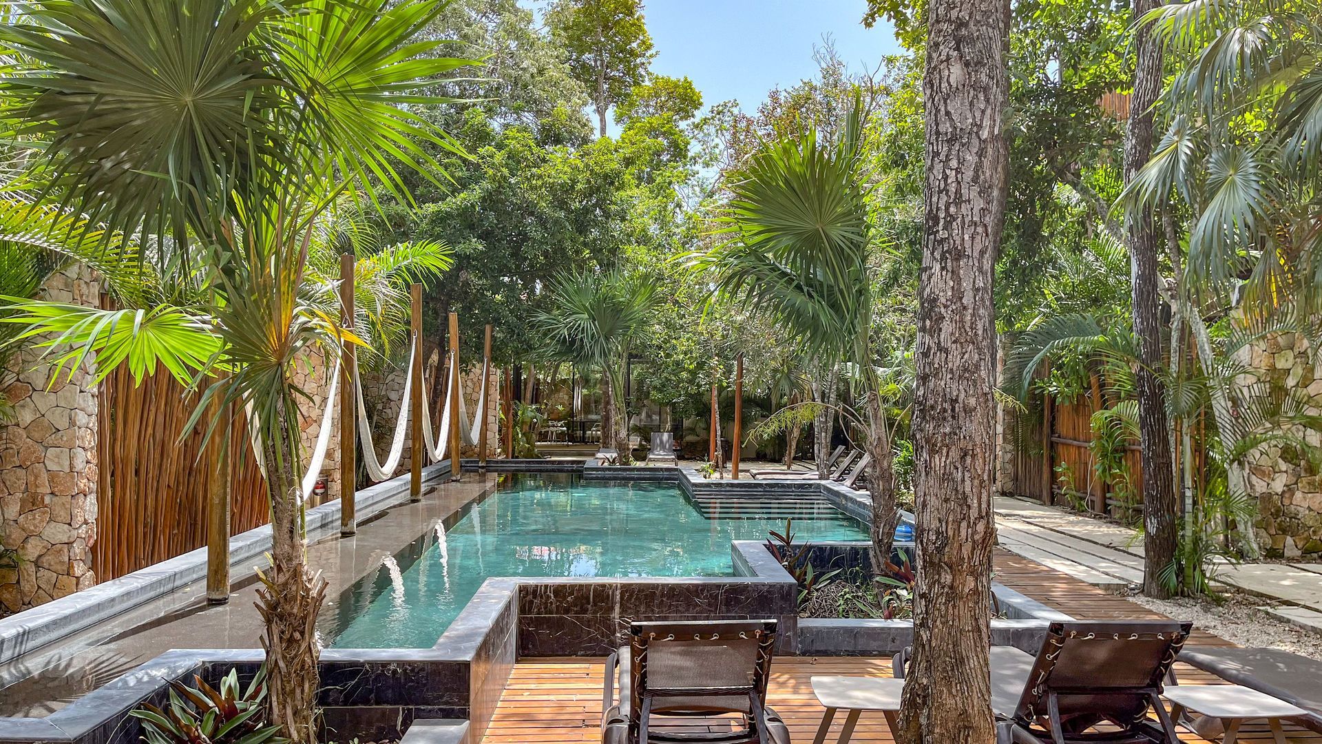 Piscina rodeada de árboles, hamacas y tumbonas. Aguas azules y un ambiente tropical.
