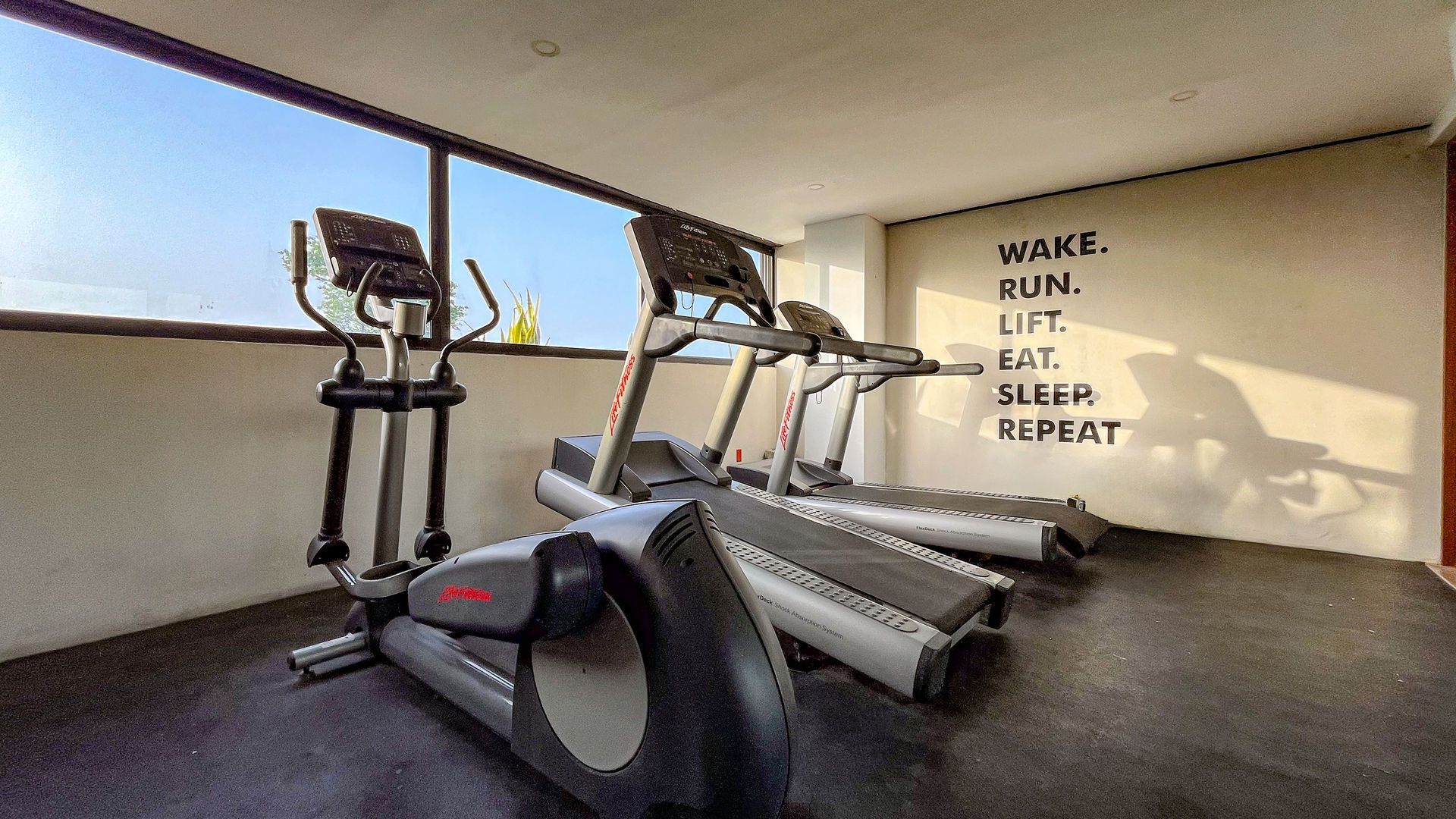 Interior de gimnasio con elíptica, cintas de correr y texto motivacional en la pared cerca de una ventana.