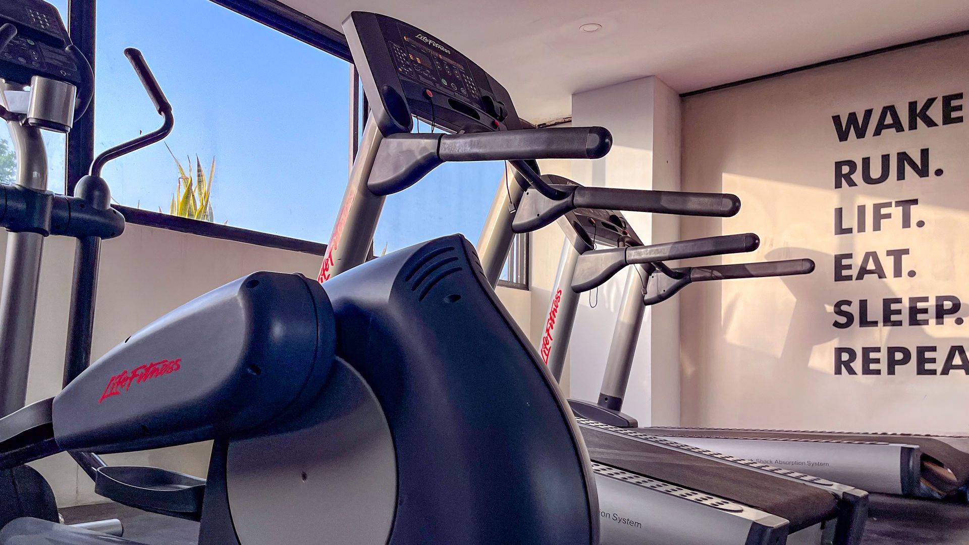 Máquina elíptica y cintas de correr en un gimnasio moderno con texto motivacional en la pared.