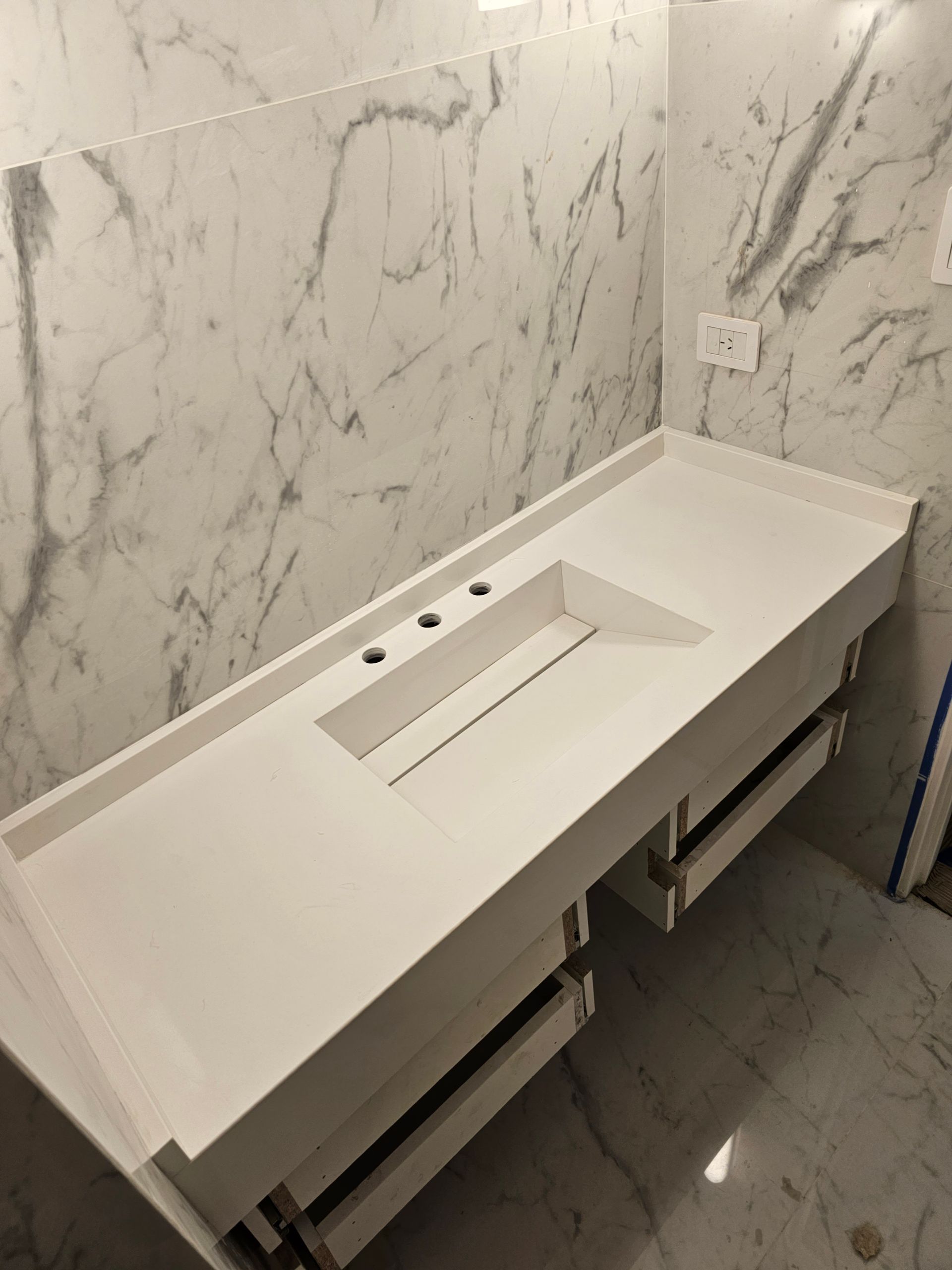 Mueble de baño blanco con lavabo integrado, tres orificios para grifería y cajones, contra pared símil mármol.