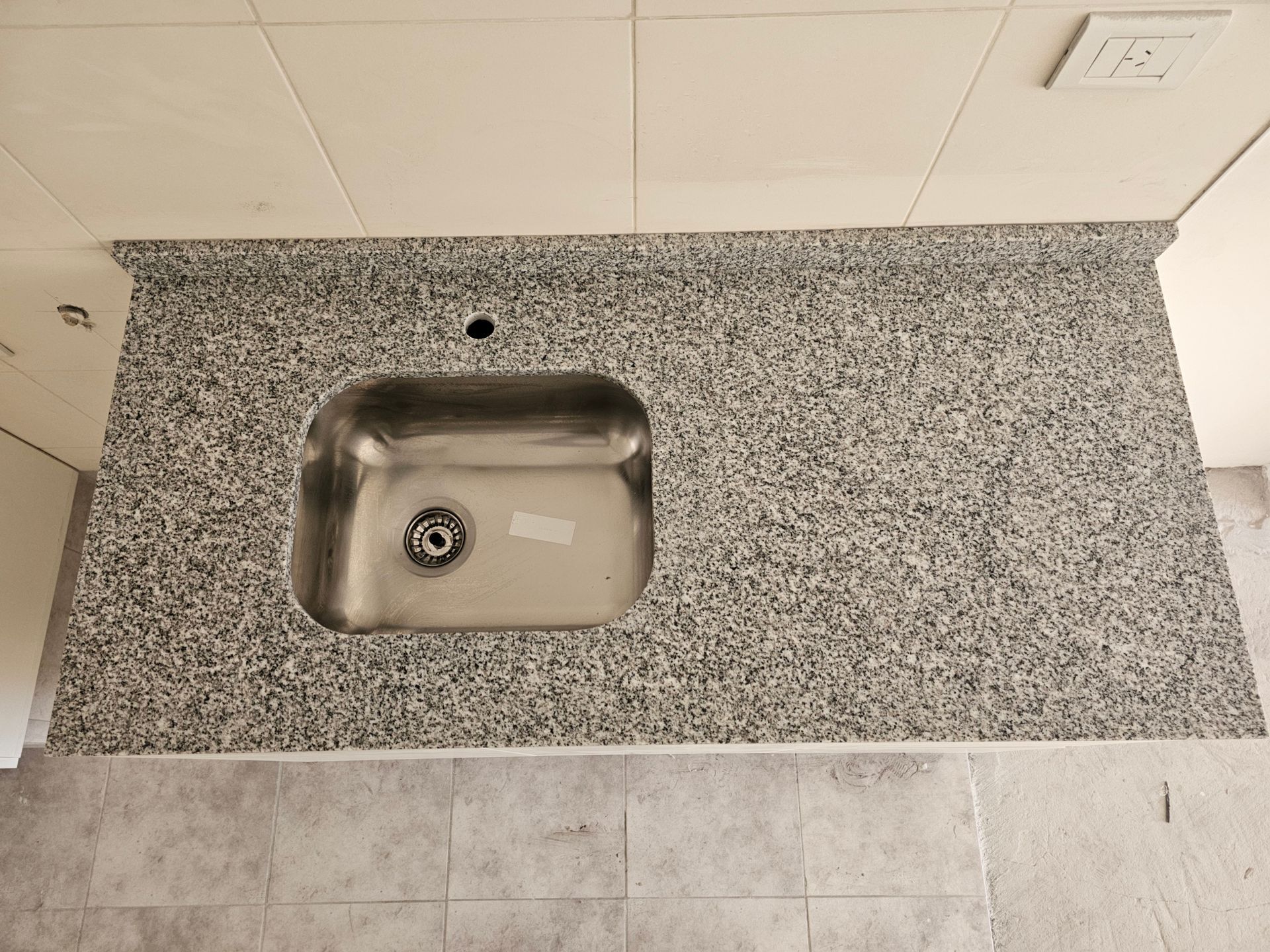Encimera de granito con fregadero de acero inoxidable, sobre un suelo de baldosas y un techo blanco.