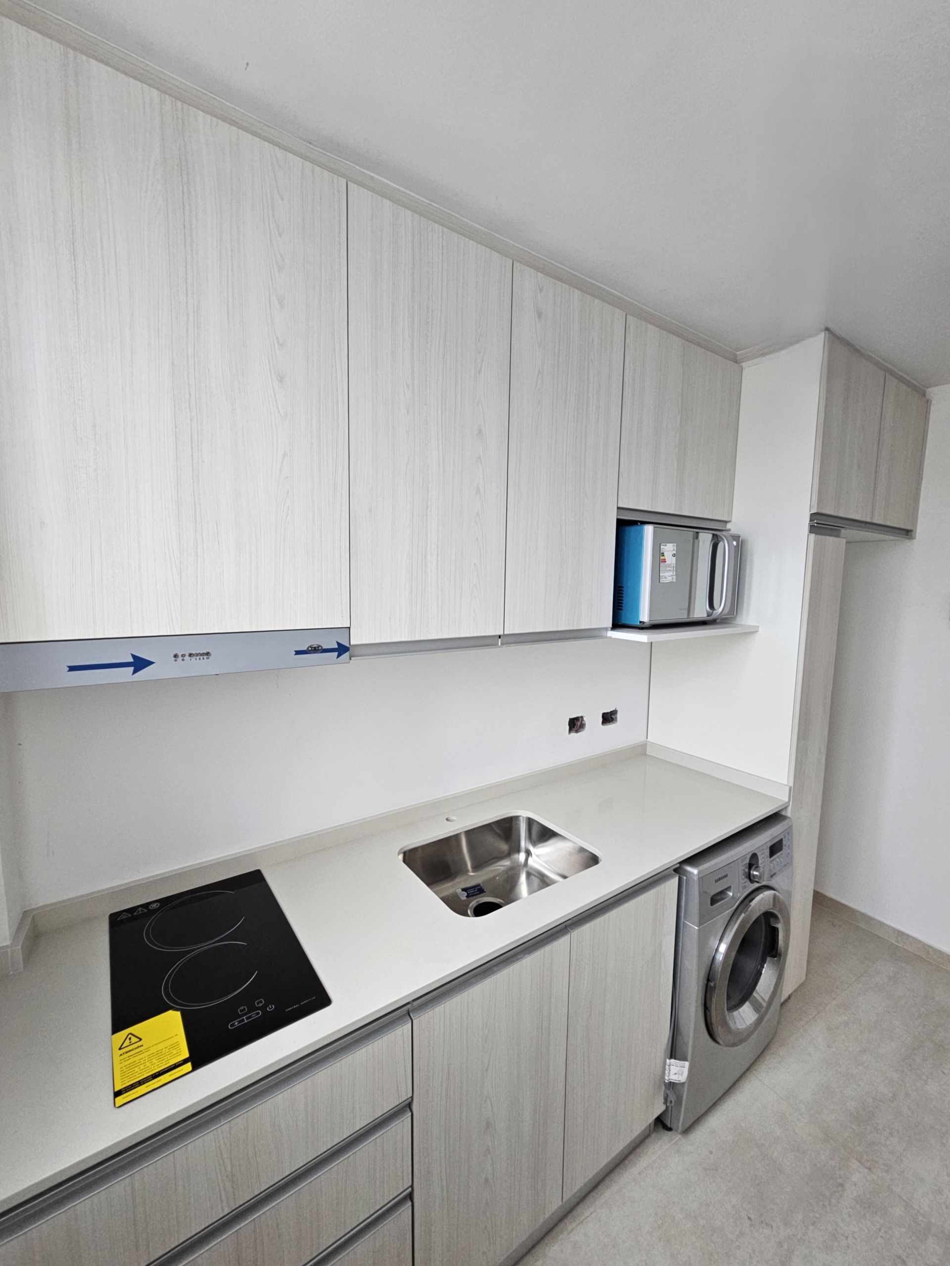 Una cocina pequeña y moderna con gabinetes de madera clara, encimeras blancas y lavadora.