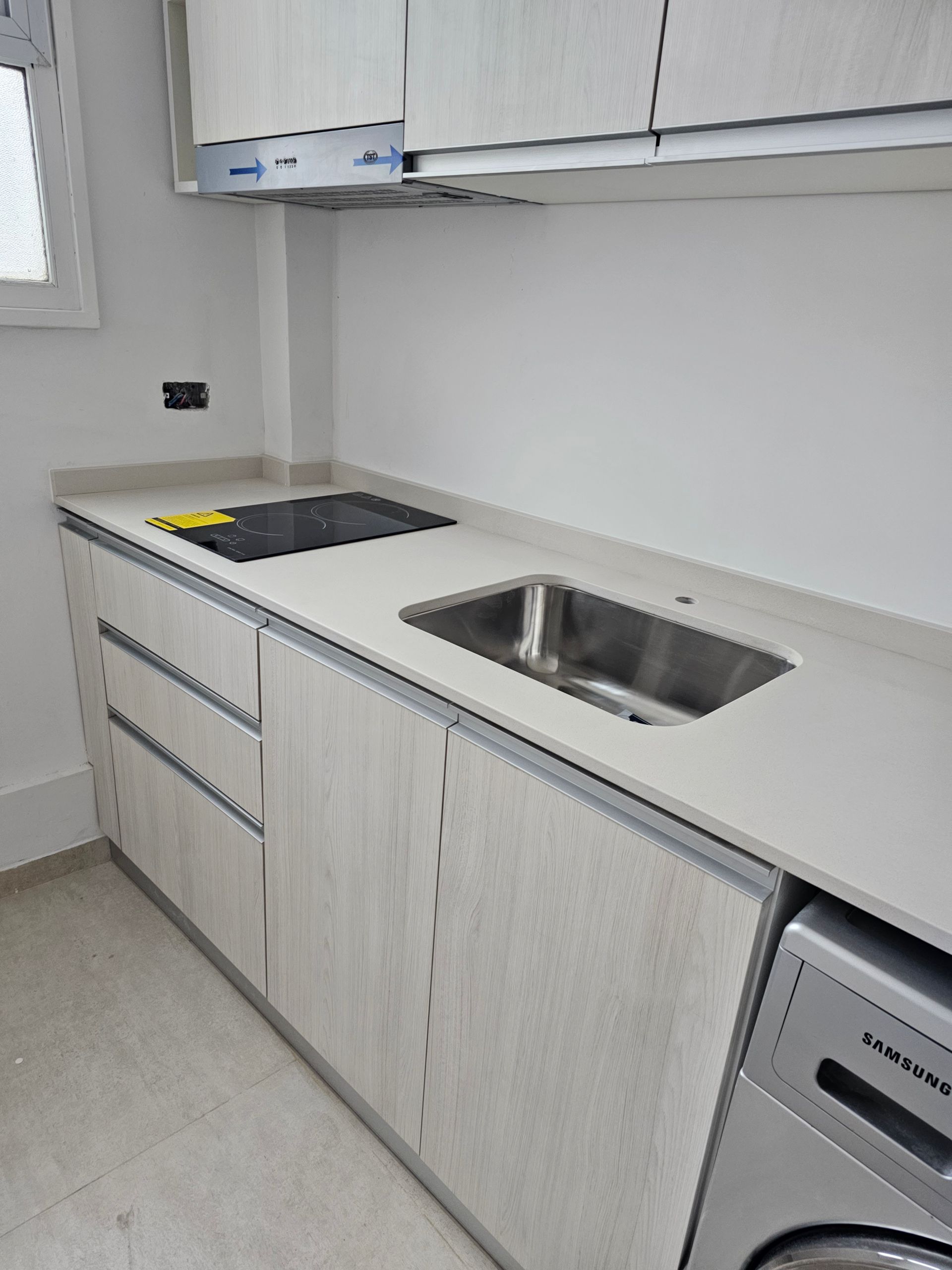 Cocina moderna con gabinetes de colores claros, fregadero de acero inoxidable, encimera y lavadora/secadora incorporadas.