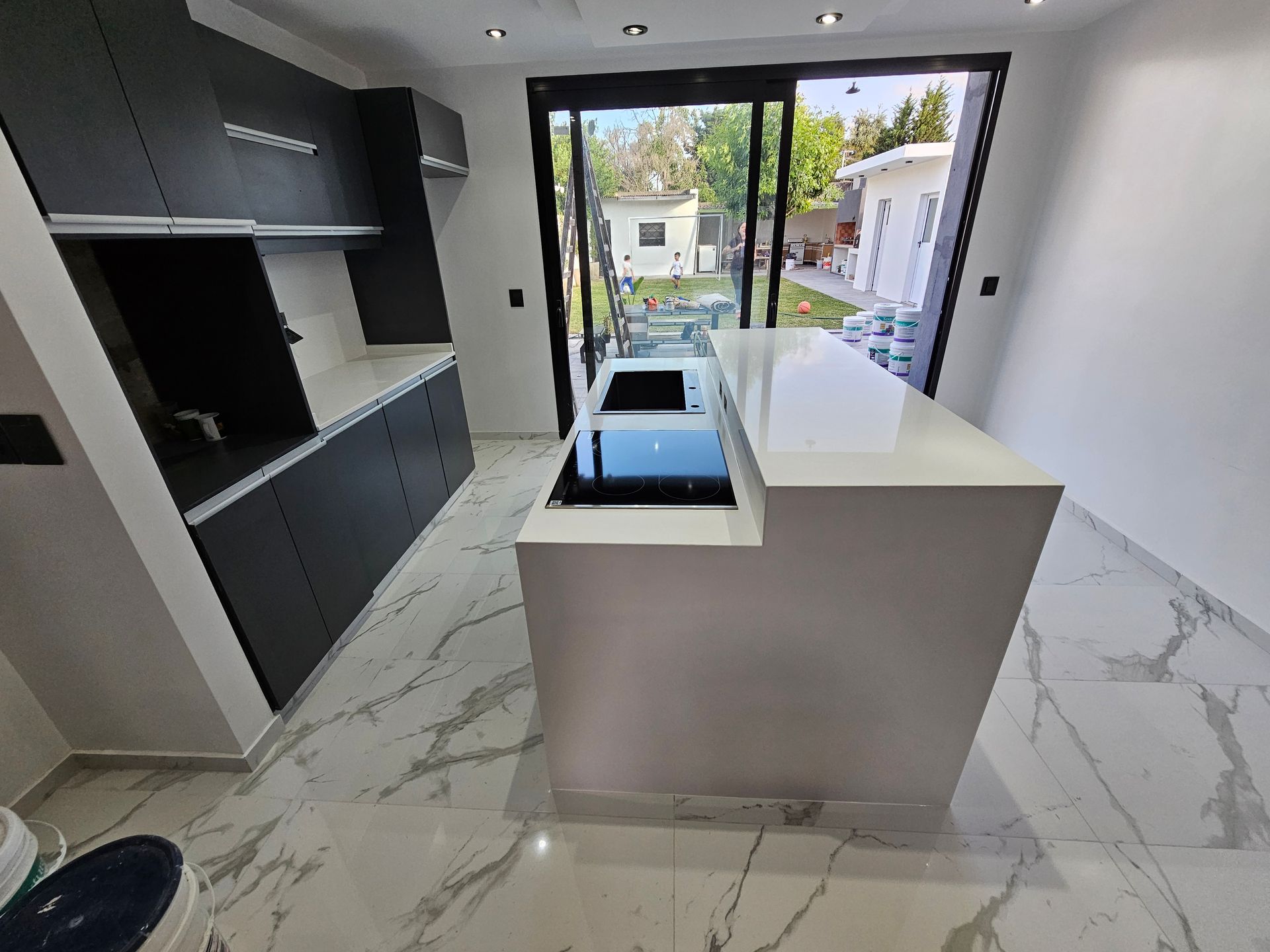 Cocina moderna con gabinetes oscuros, encimeras blancas y una isla con anafe, con vista a través de puertas de vidrio.