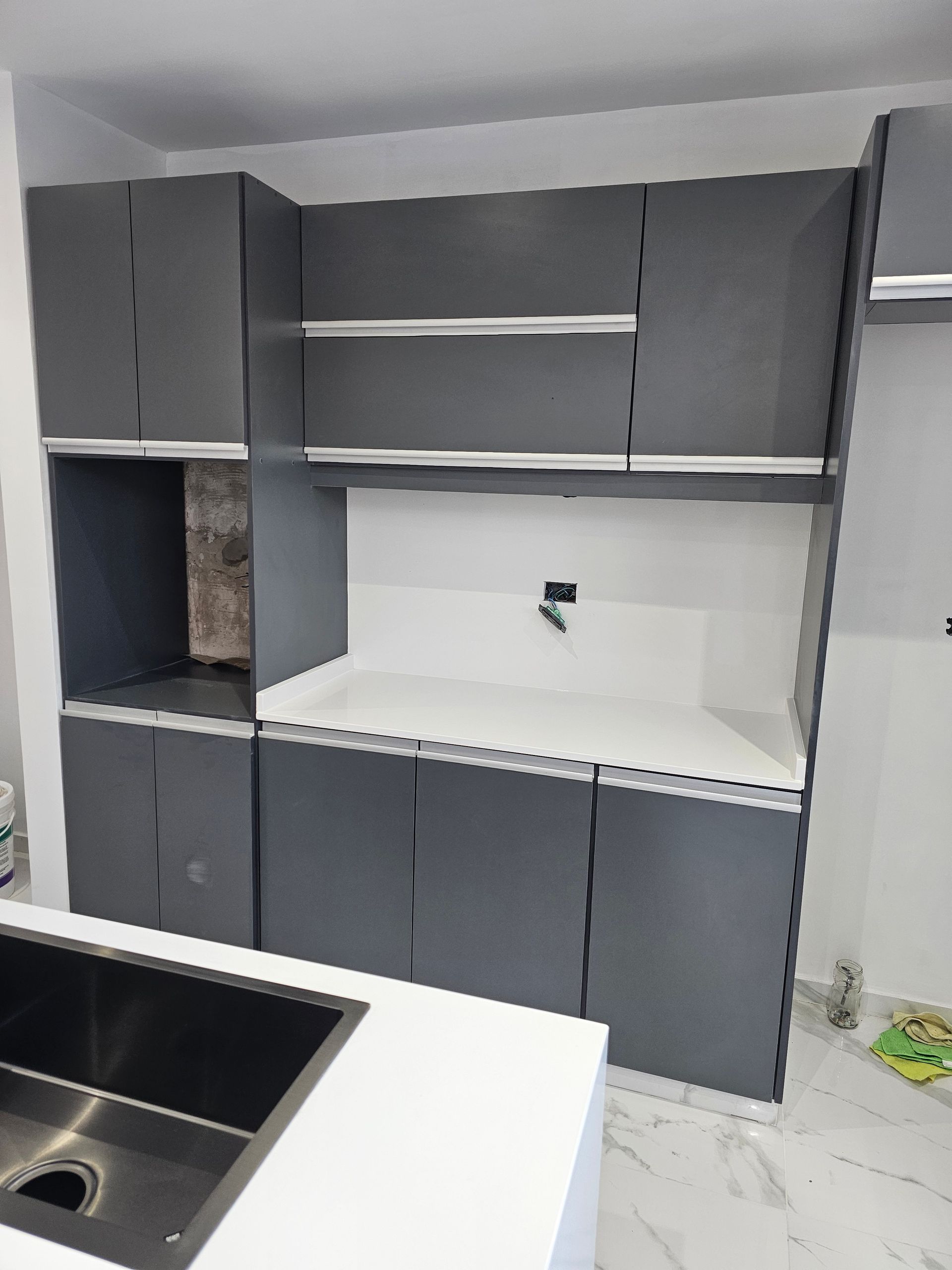 Muebles de cocina grises con horno empotrado, estante abierto y encimera blanca. Un fregadero de acero inoxidable