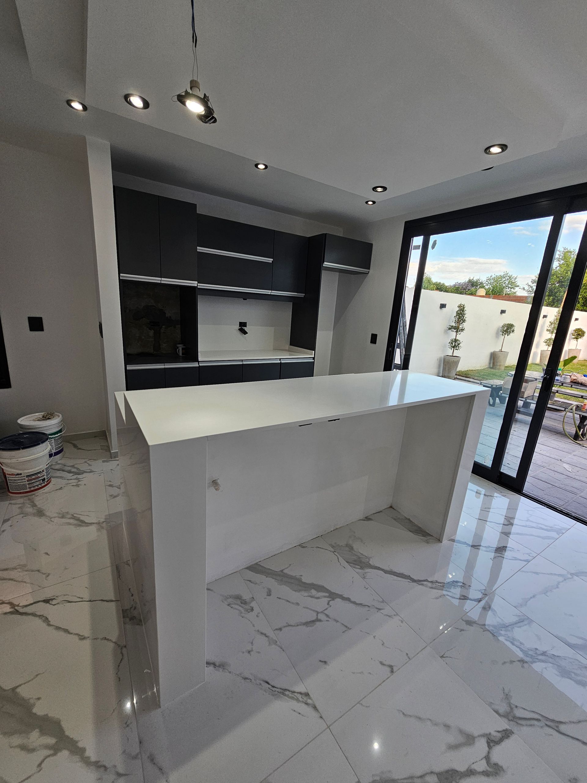 Cocina moderna blanca con isla, gabinetes grises y piso de mármol, junto a puertas de vidrio.