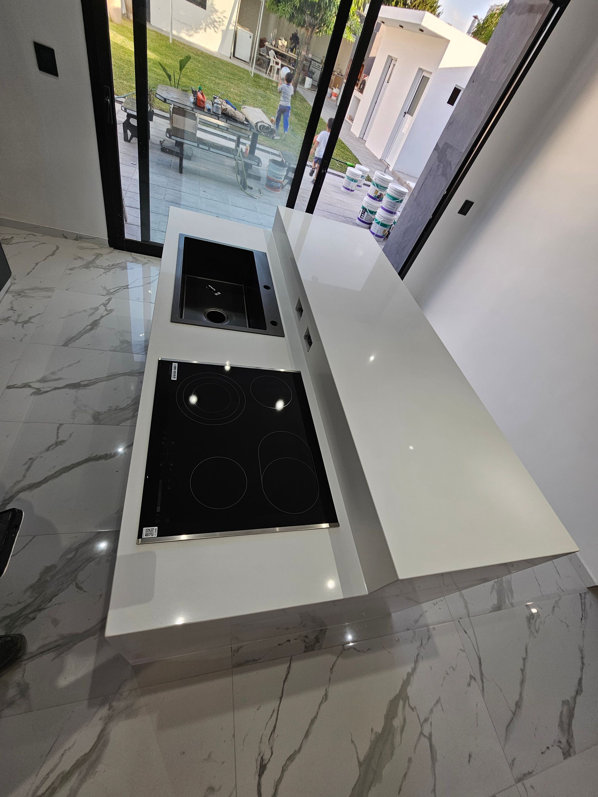 Isla de cocina blanca con anafe y fregadero, frente a grandes ventanales con vista exterior.