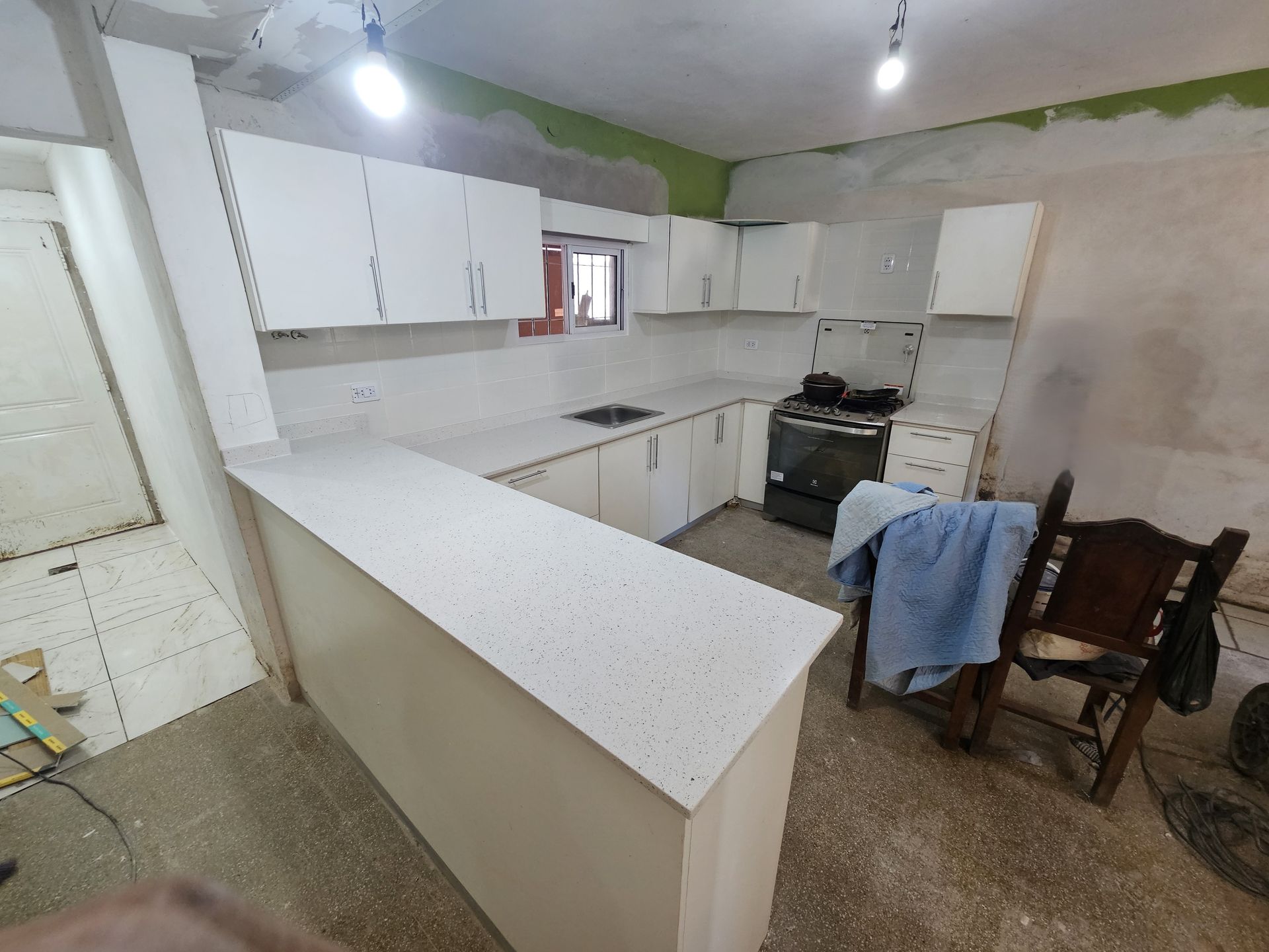 Gabinetes y encimera blancos en una cocina en construcción. Se ven una estufa negra y una silla.