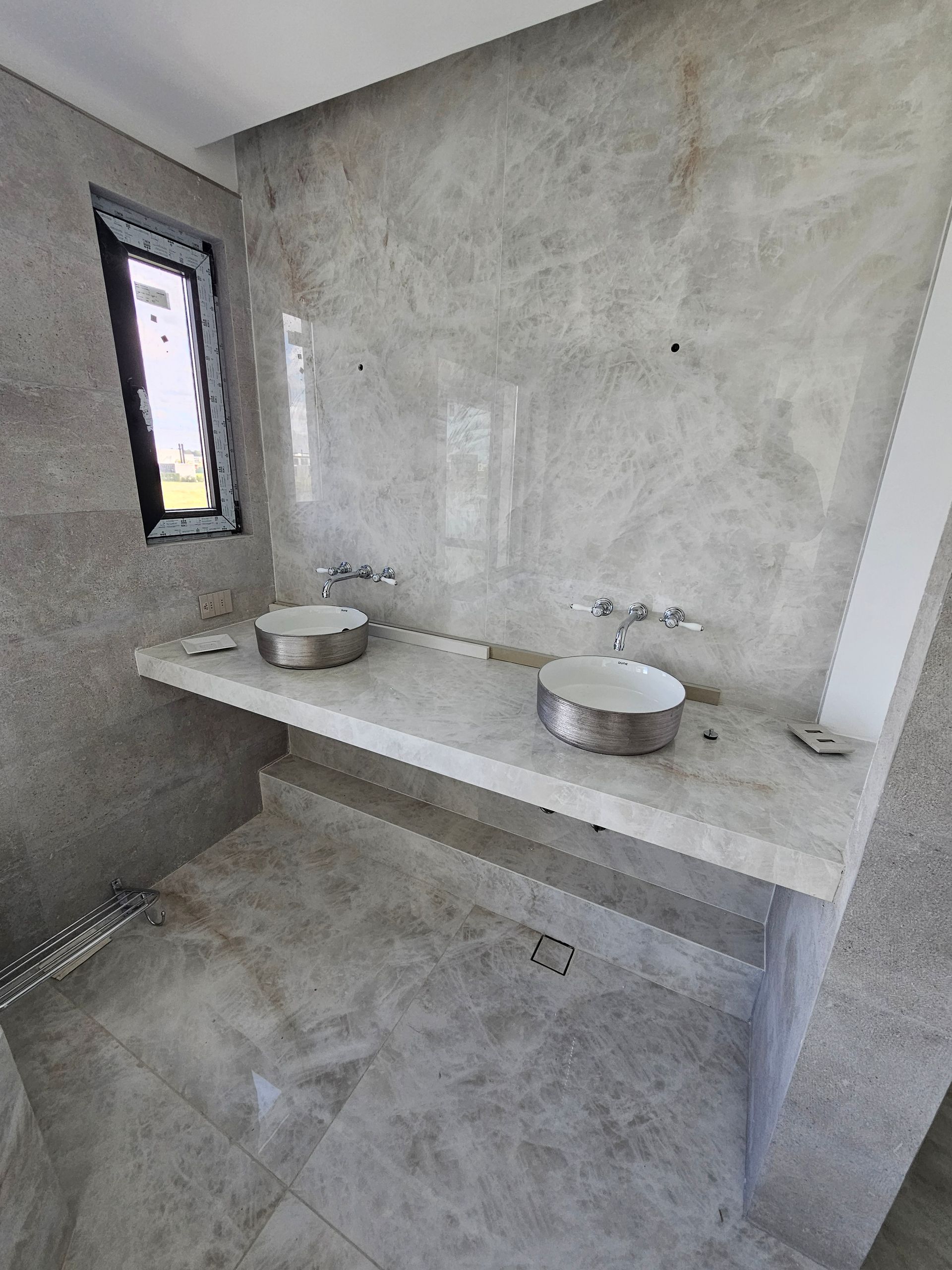 Baño con dos lavabos plateados sobre una encimera de mármol contra una pared revestida de mármol.