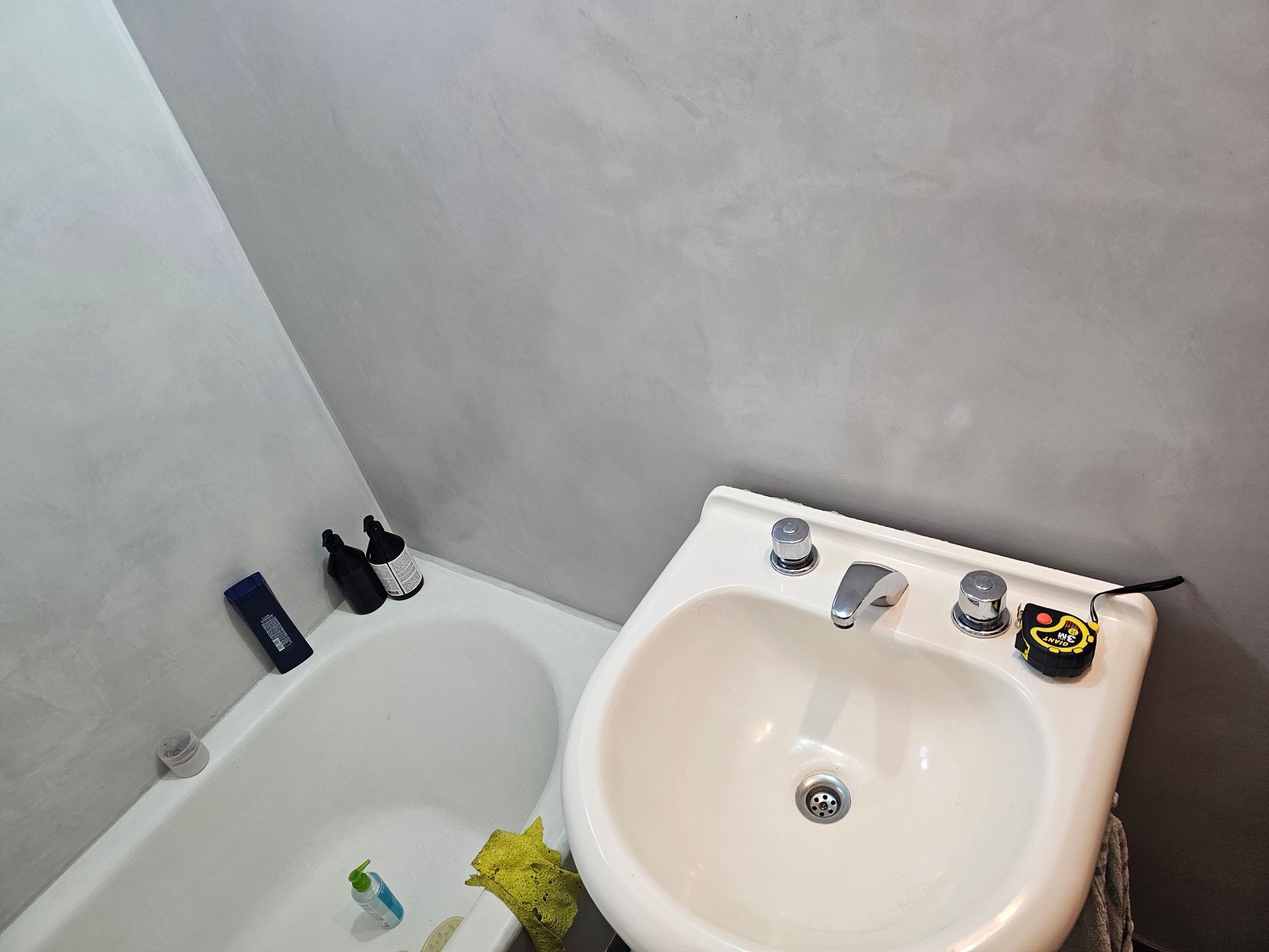 Baño con lavabo blanco, bañera y paredes grises. Botellas en el borde de la bañera.