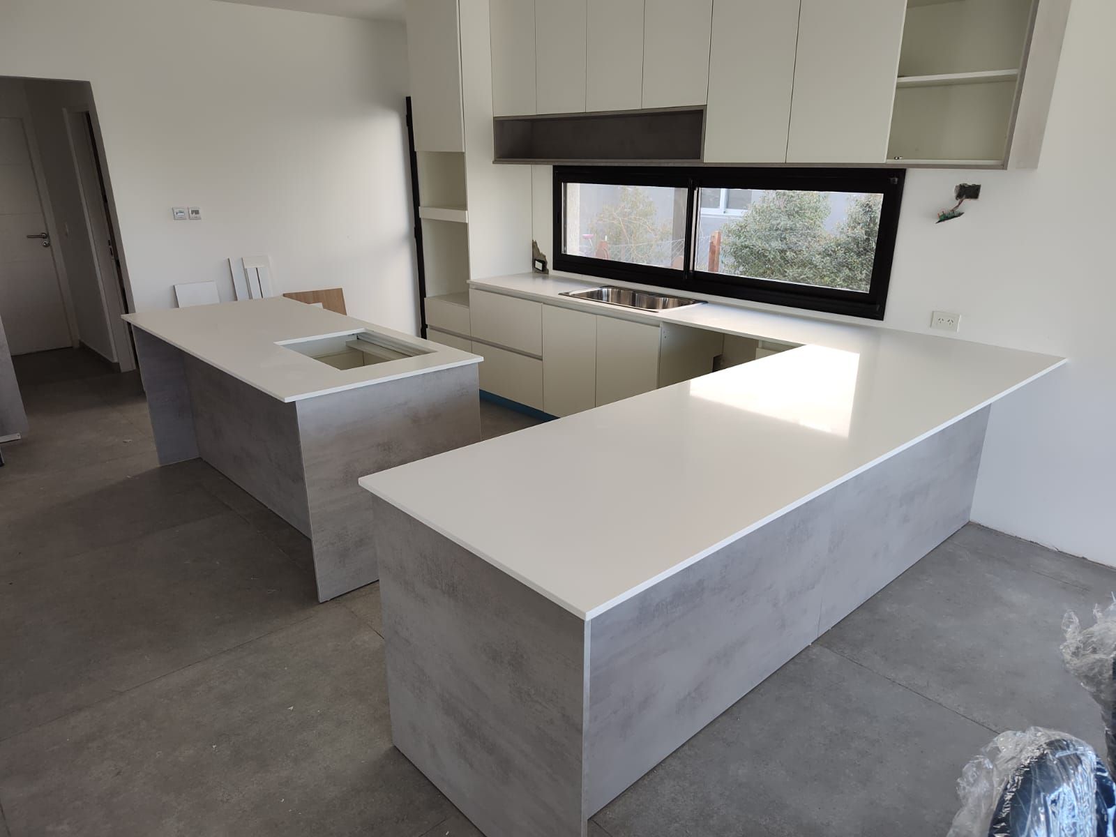 Cocina moderna con islas de hormigón gris y encimeras blancas, gabinetes al fondo.