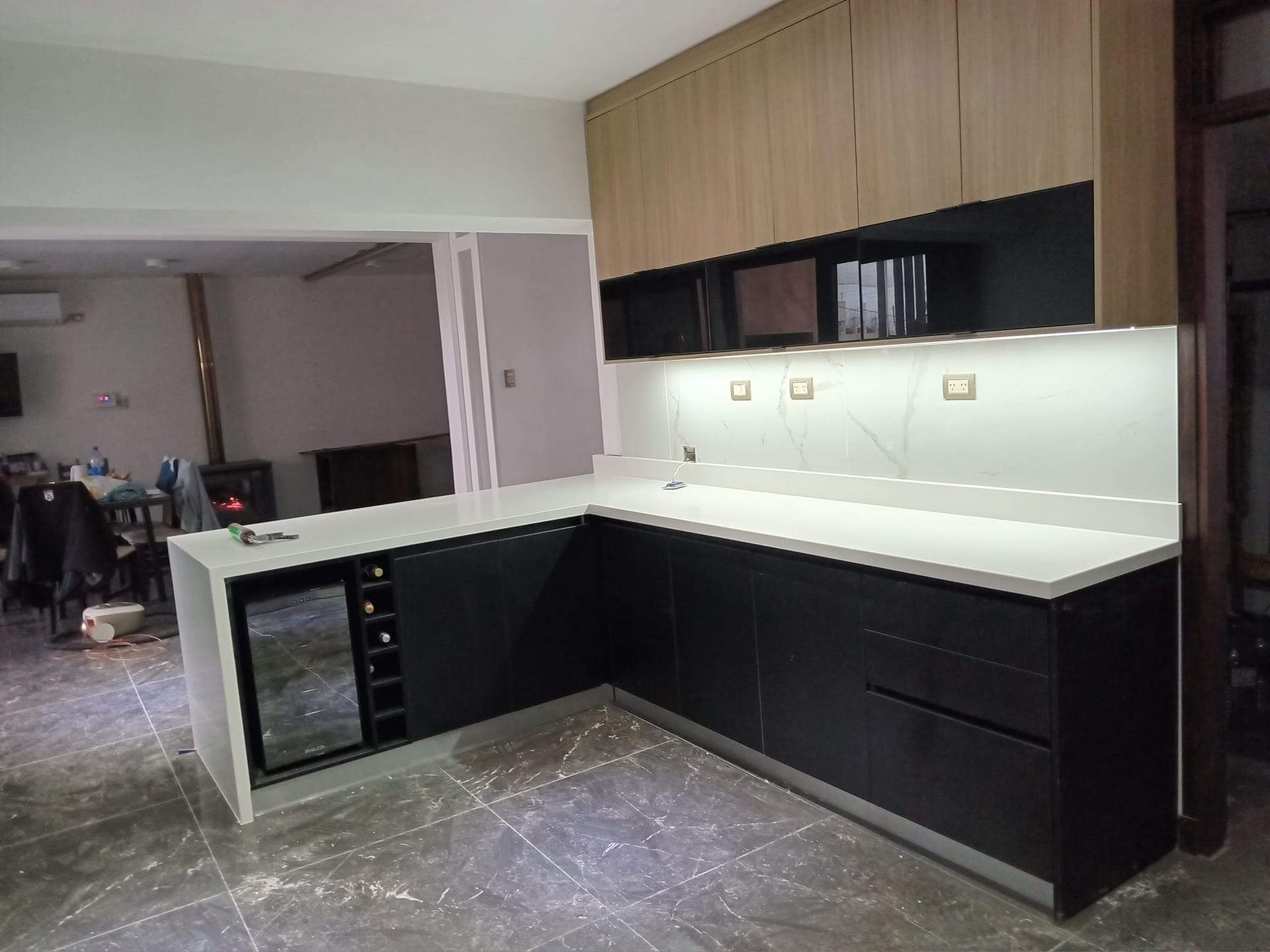 Cocina moderna con gabinetes negros, encimera blanca, refrigerador para vinos y gabinetes superiores en tono madera.