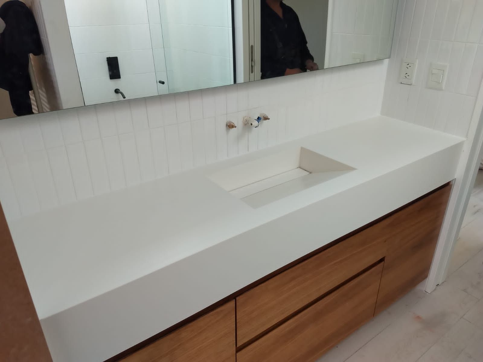 Encimera blanca con lavabo empotrado sobre armarios de madera en un baño. Hay un espejo encima.