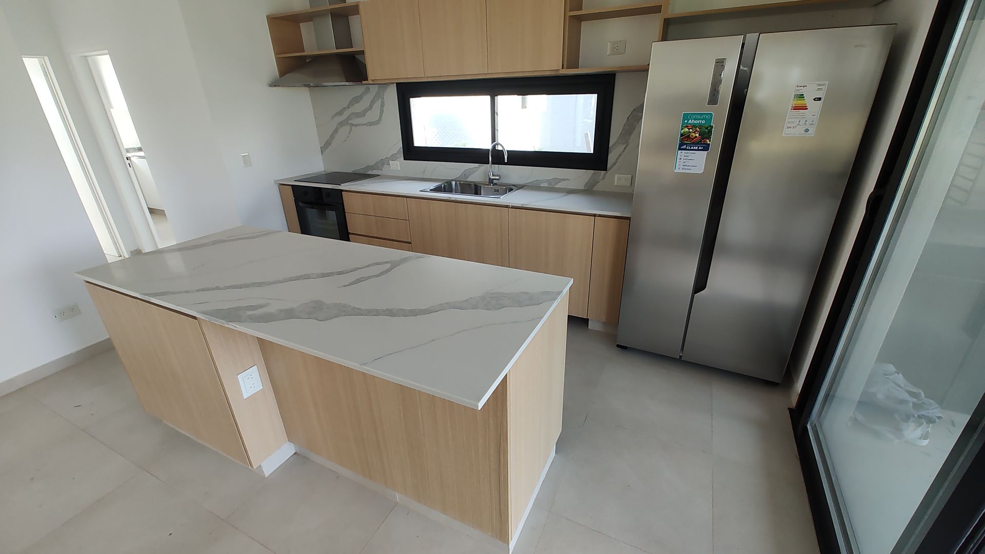 Cocina moderna con gabinetes de madera clara, refrigerador de acero inoxidable y una isla con encimera de mármol.