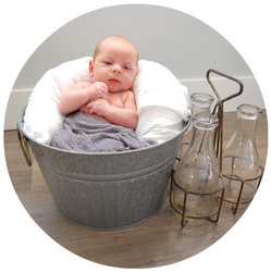 Baby resting in a metal tub with white pillows, beside two glass bottles on a wooden floor.