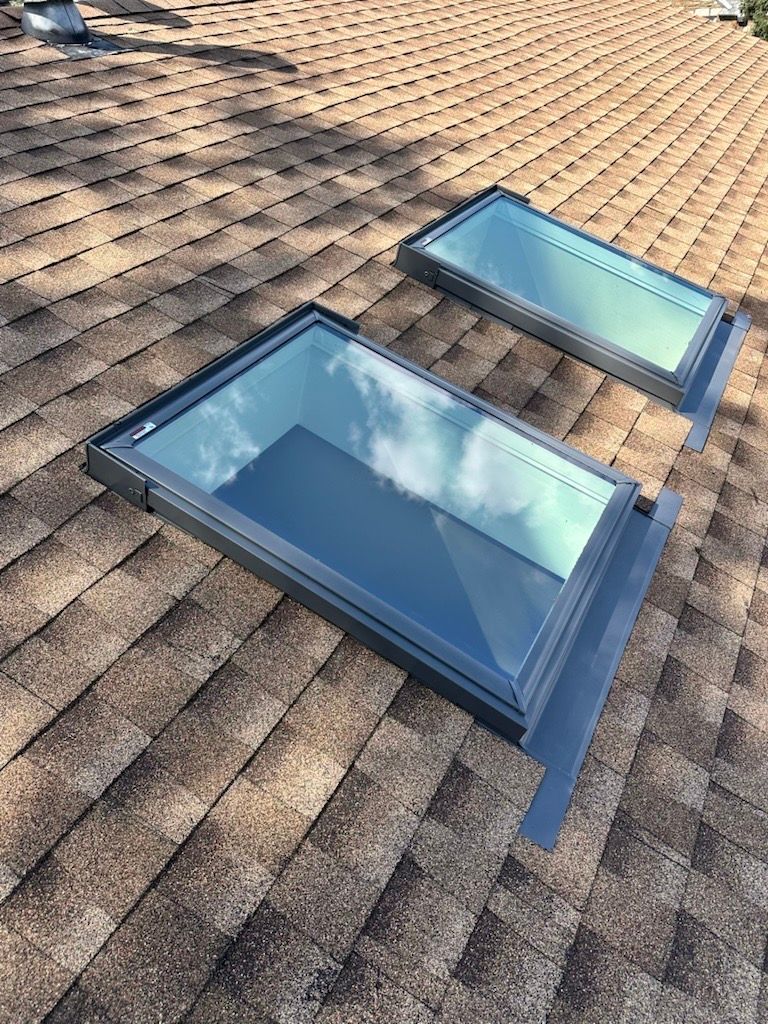 Two rectangular skylights on a brown shingled roof reflect a cloudy sky.