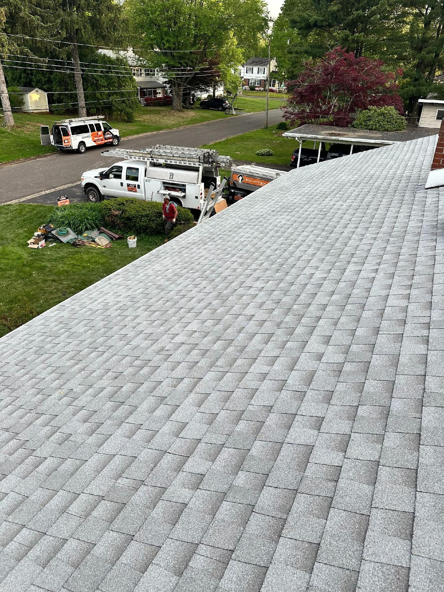 Gray shingle roof with workers and trucks in a residential area; roofing installation in progress.