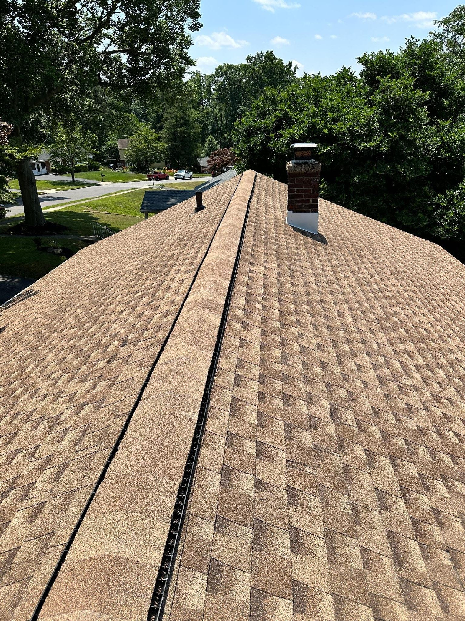 View of a brown shingle roof with a central ridge and chimney, trees in the background.