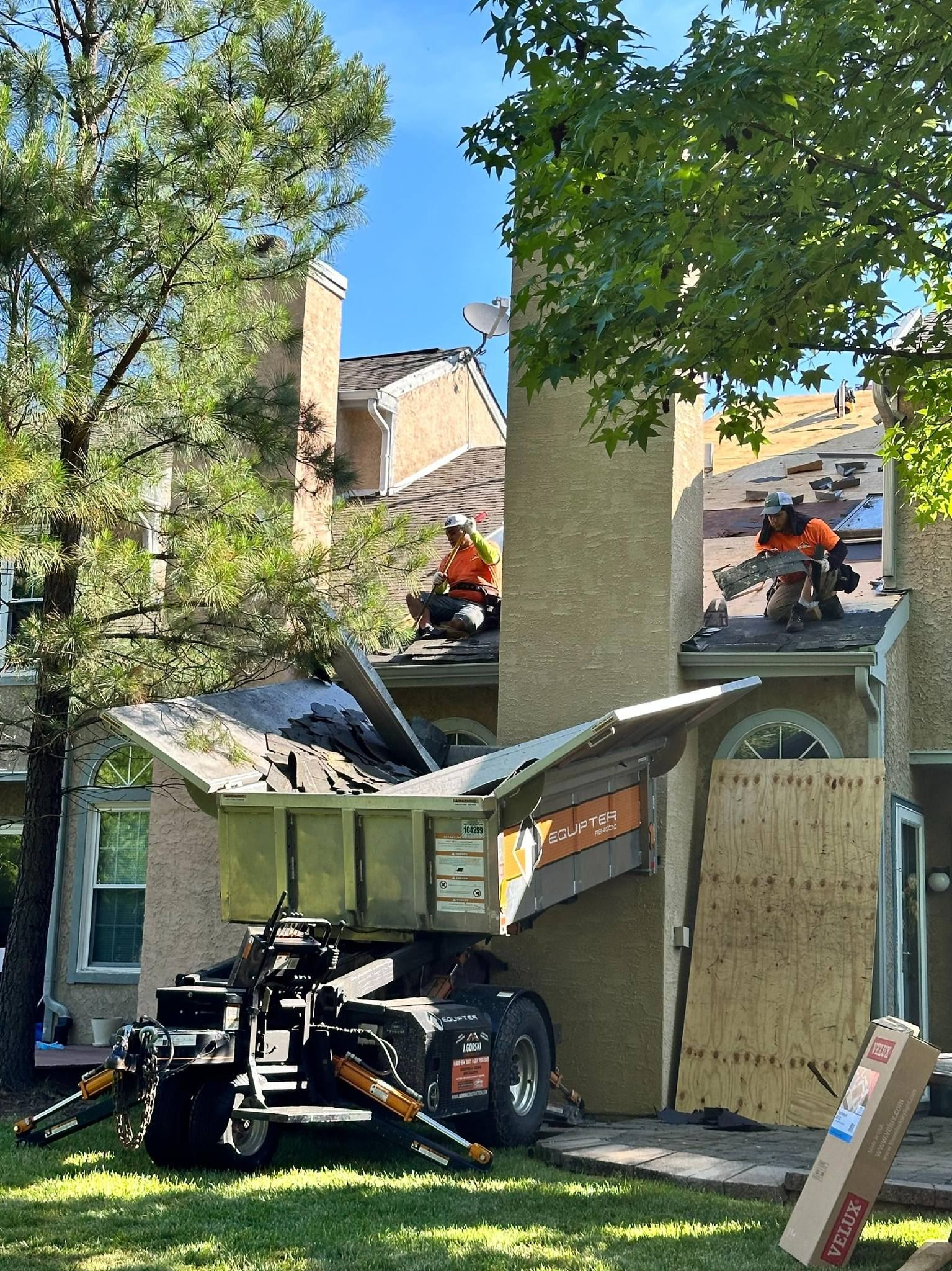 Roofers replacing shingles on a house; debris removal in progress with machinery, under a blue sky.