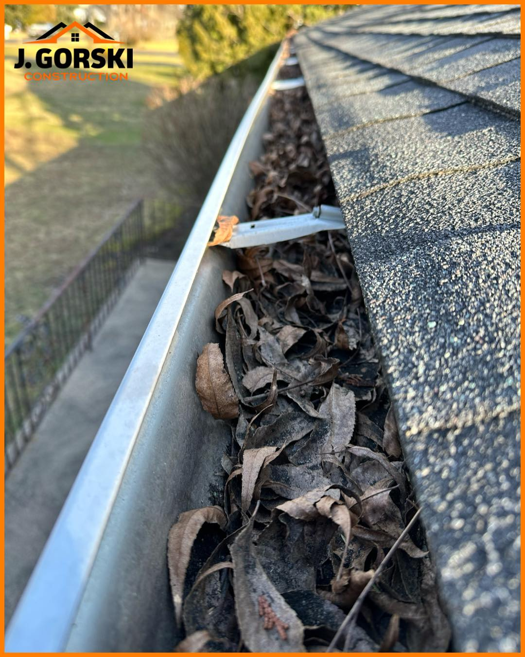 Gutter overflowing with dead leaves next to a dark shingle roof. Sunny outdoor setting.