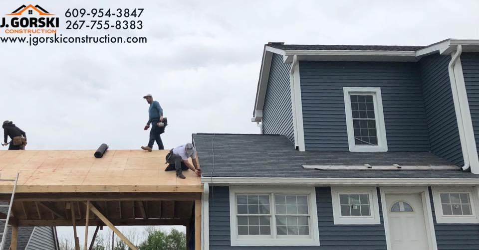 Construction workers on a roof, installing shingles. Blue siding building and wooden supports, under overcast sky.