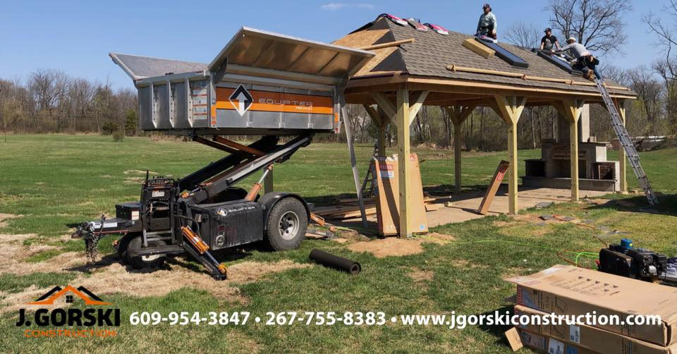 Construction workers roofing a wooden pavilion with a lift and materials outdoors.