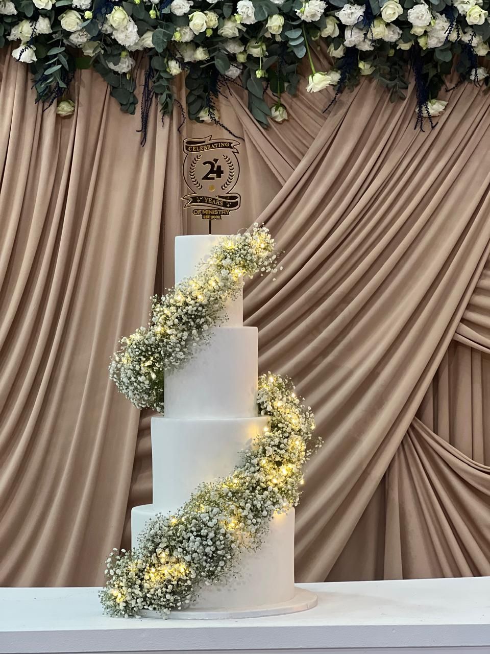 Four-tiered white cake with flowers and string lights. Decorated for a celebration, in front of draped beige fabric.