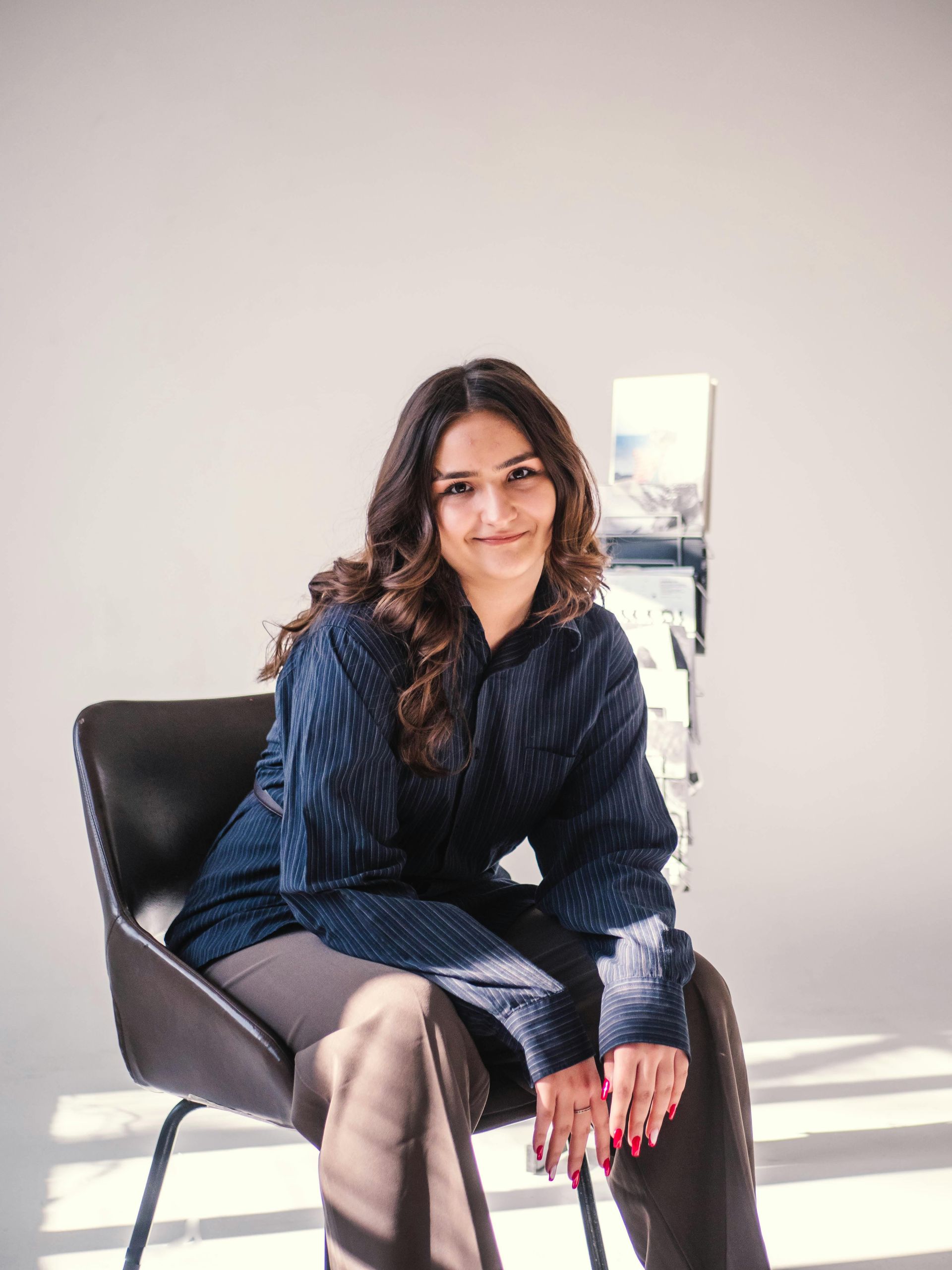 A person with long, wavy brown hair sits in a dark chair, wearing a textured navy shirt and brown trousers.