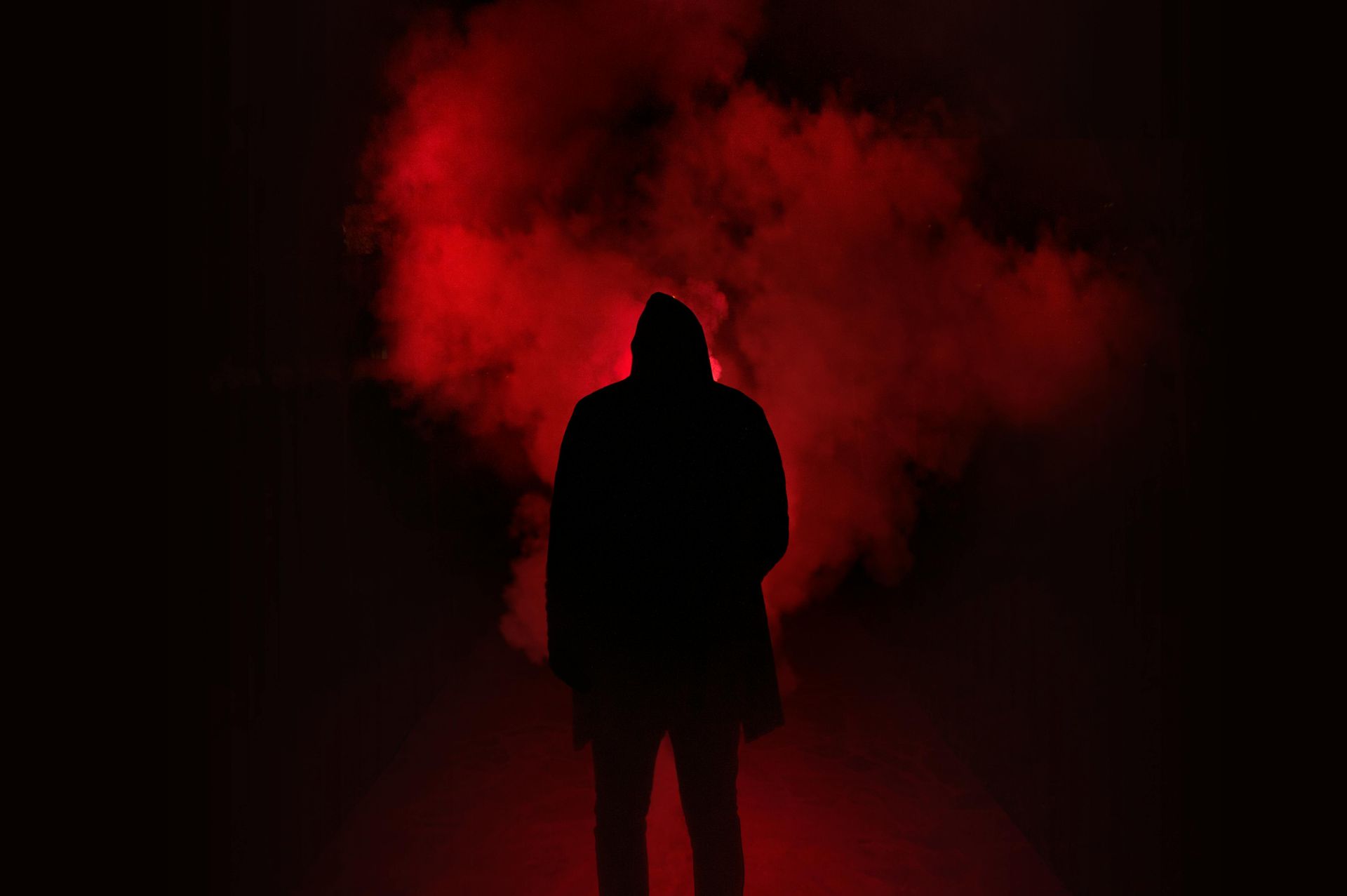 A silhouette of a man in a hood standing in a dark room surrounded by red smoke.